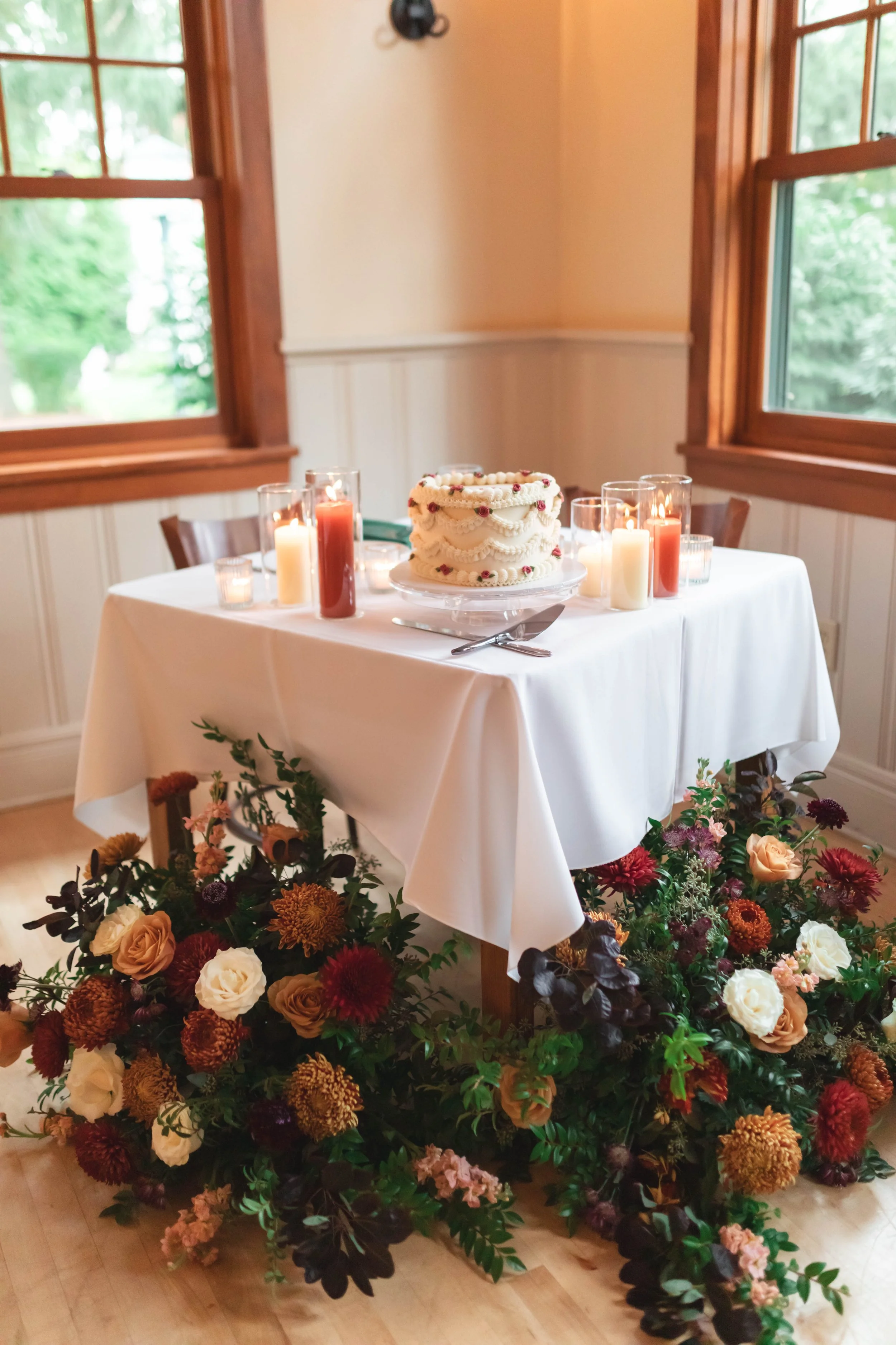 A table with a white tablecloth holding a decorated cake with icing and small flowers, topped with candles in glass holders of various heights, and surrounded by floral arrangements of mixed flowers and greenery.