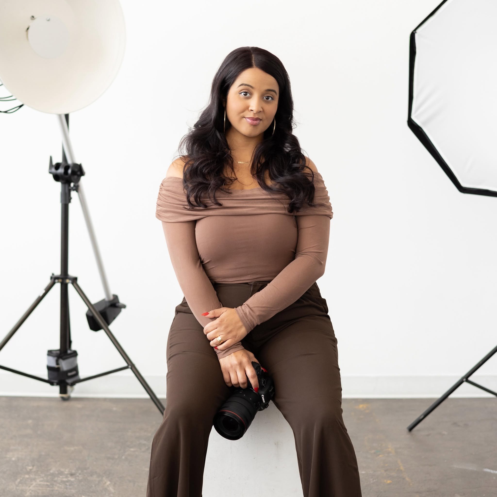 A woman with long dark hair wearing a brown long sleeve top and brown pants, sitting outdoors with photography equipment around her, including a camera on a lens and studio lights, against a white background.
