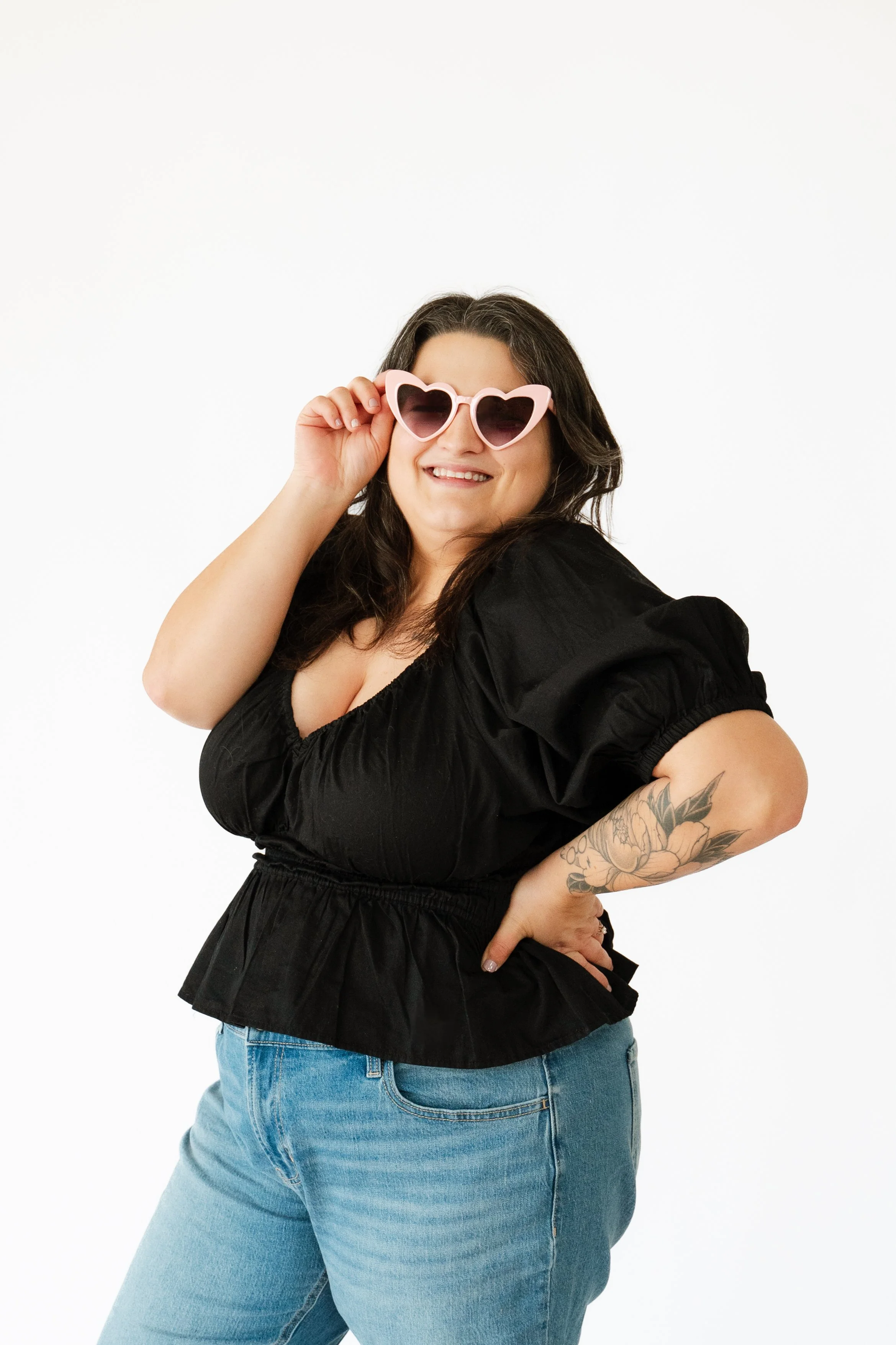 A woman smiling and wearing heart-shaped pink sunglasses, a black top, and blue jeans, standing against a white background.