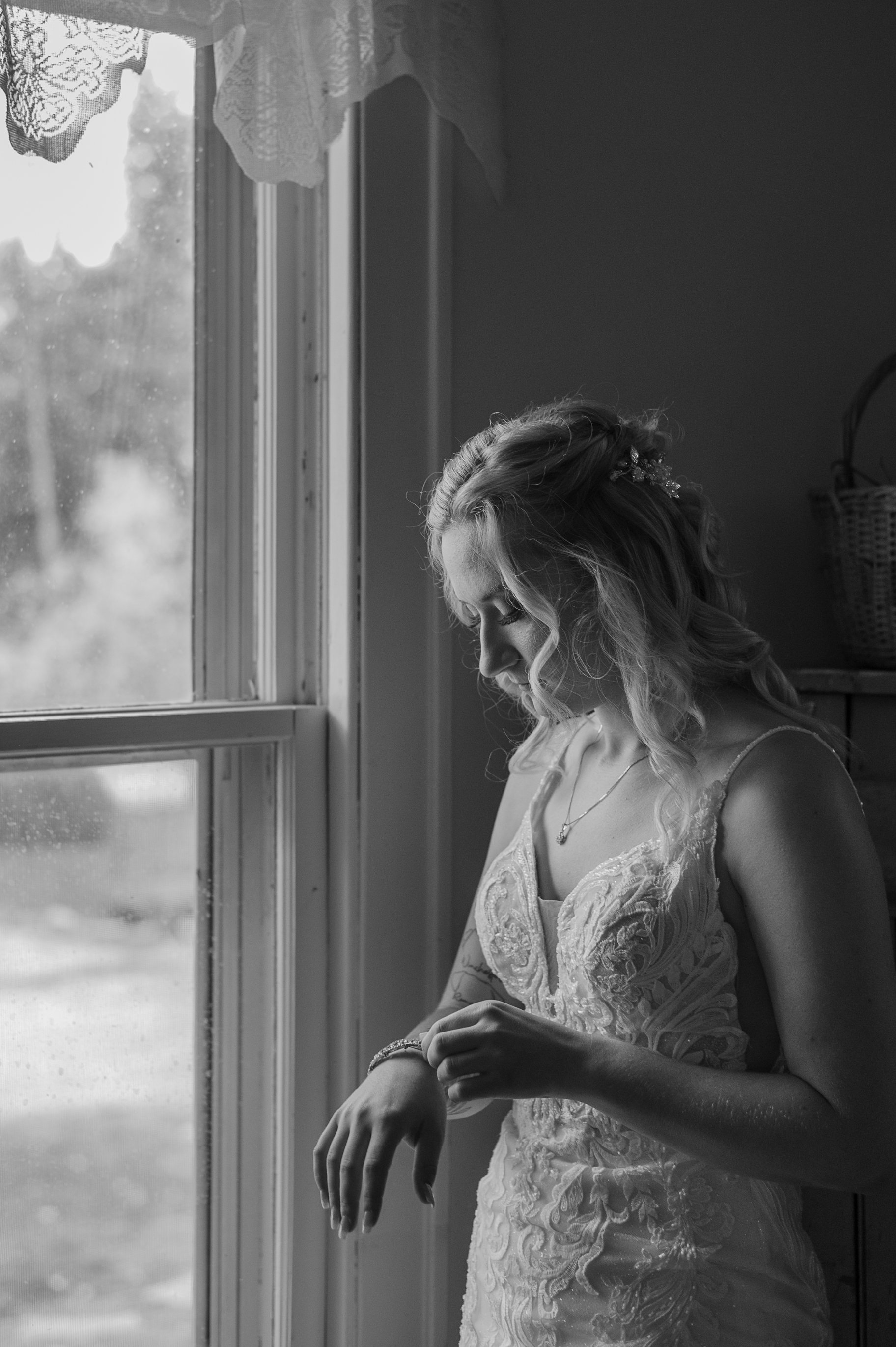 A woman in a lace wedding dress standing by a window, looking down and adjusting her bracelet.