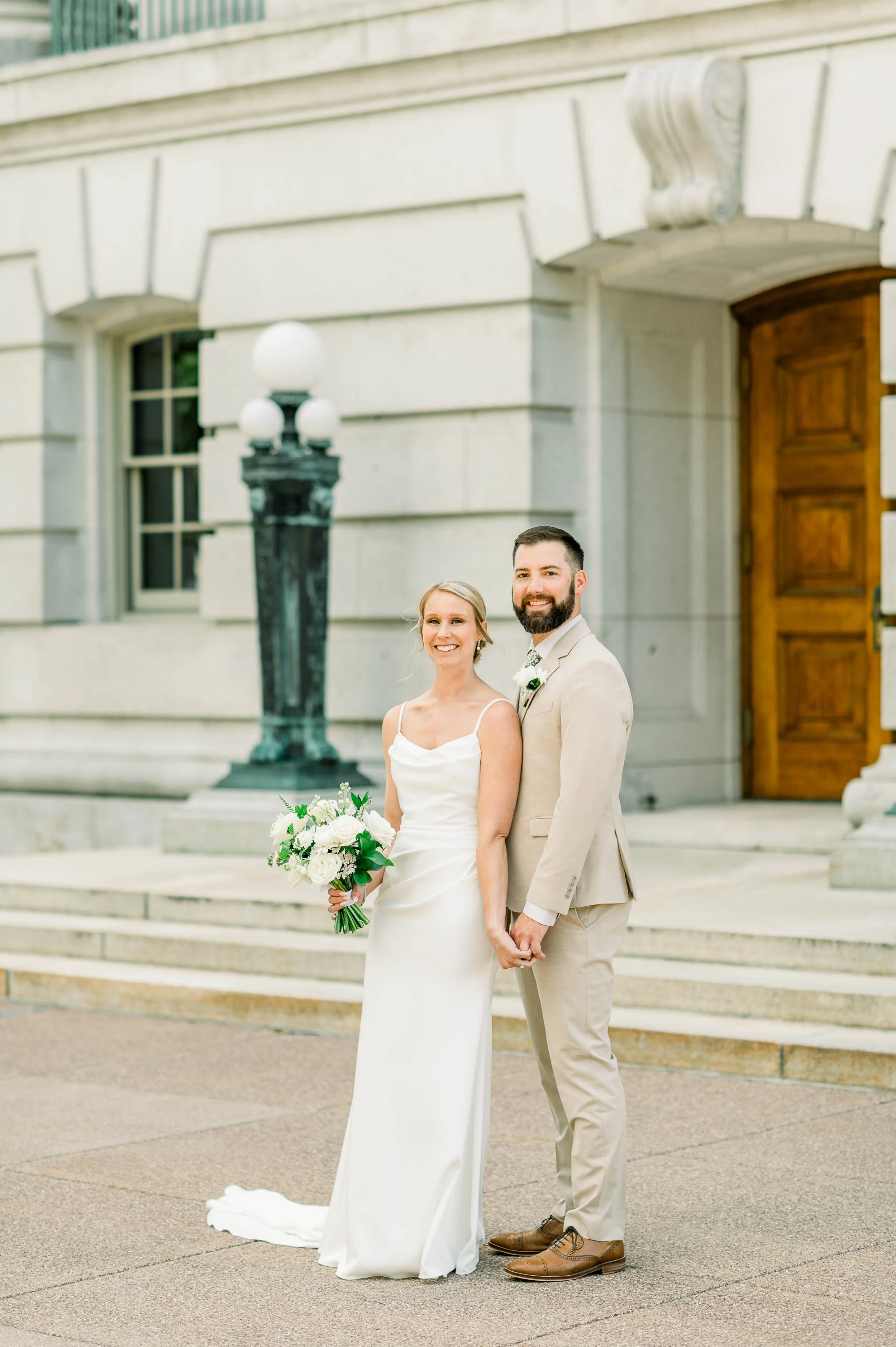 A bride and groom standing hand in hand on the steps of a historic building, smiling for a wedding photo, with a black lamppost and a large wooden door in the background.