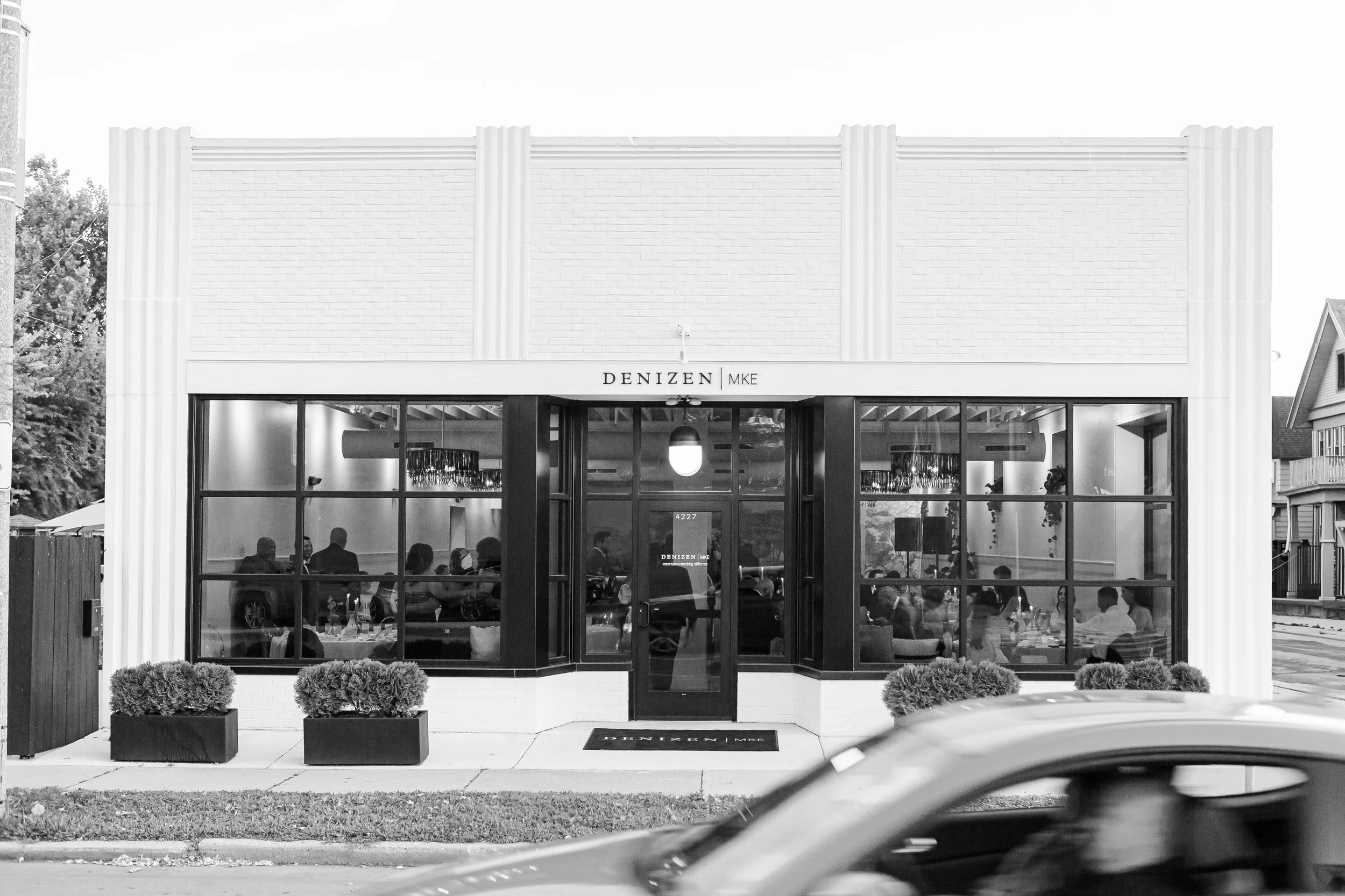 Front view of a restaurant with large glass windows, a sign above the door reading 'DENIZEN MKE,' and several patrons visible inside. Small bushes are in planters outside, and a part of a car is in the foreground.