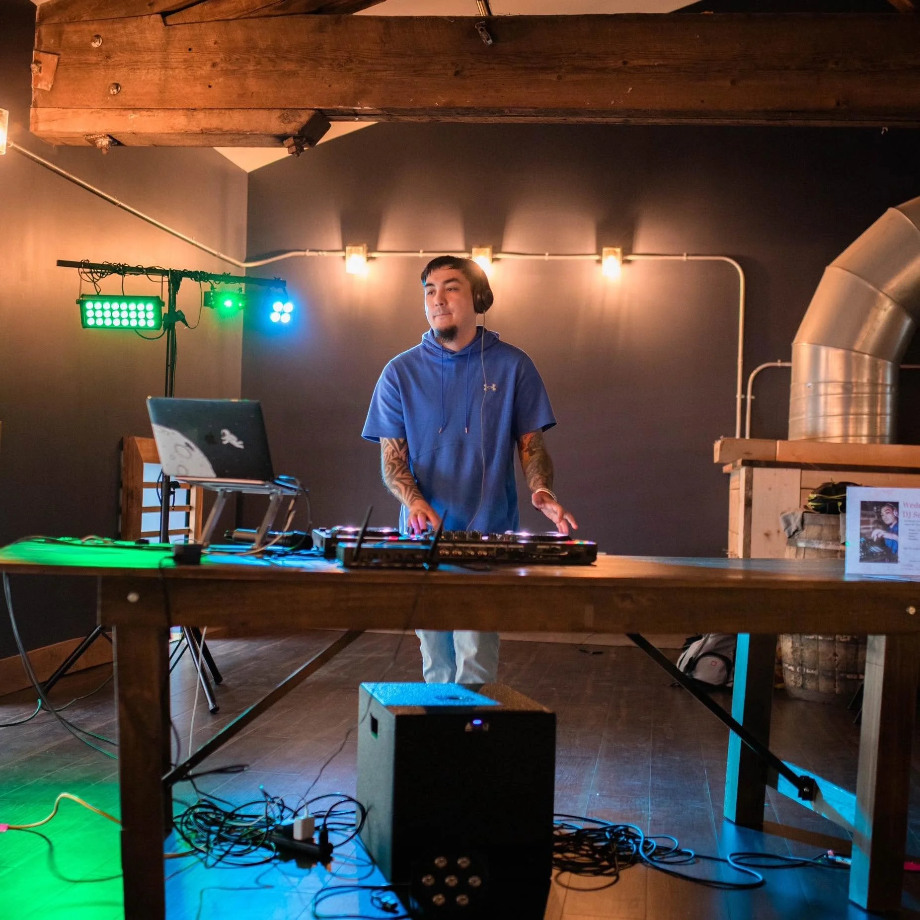 A DJ wearing a blue hoodie and headphones standing behind DJ equipment in a dimly lit room with industrial style pipes and wooden beams. Colored lights are shining on the setup.