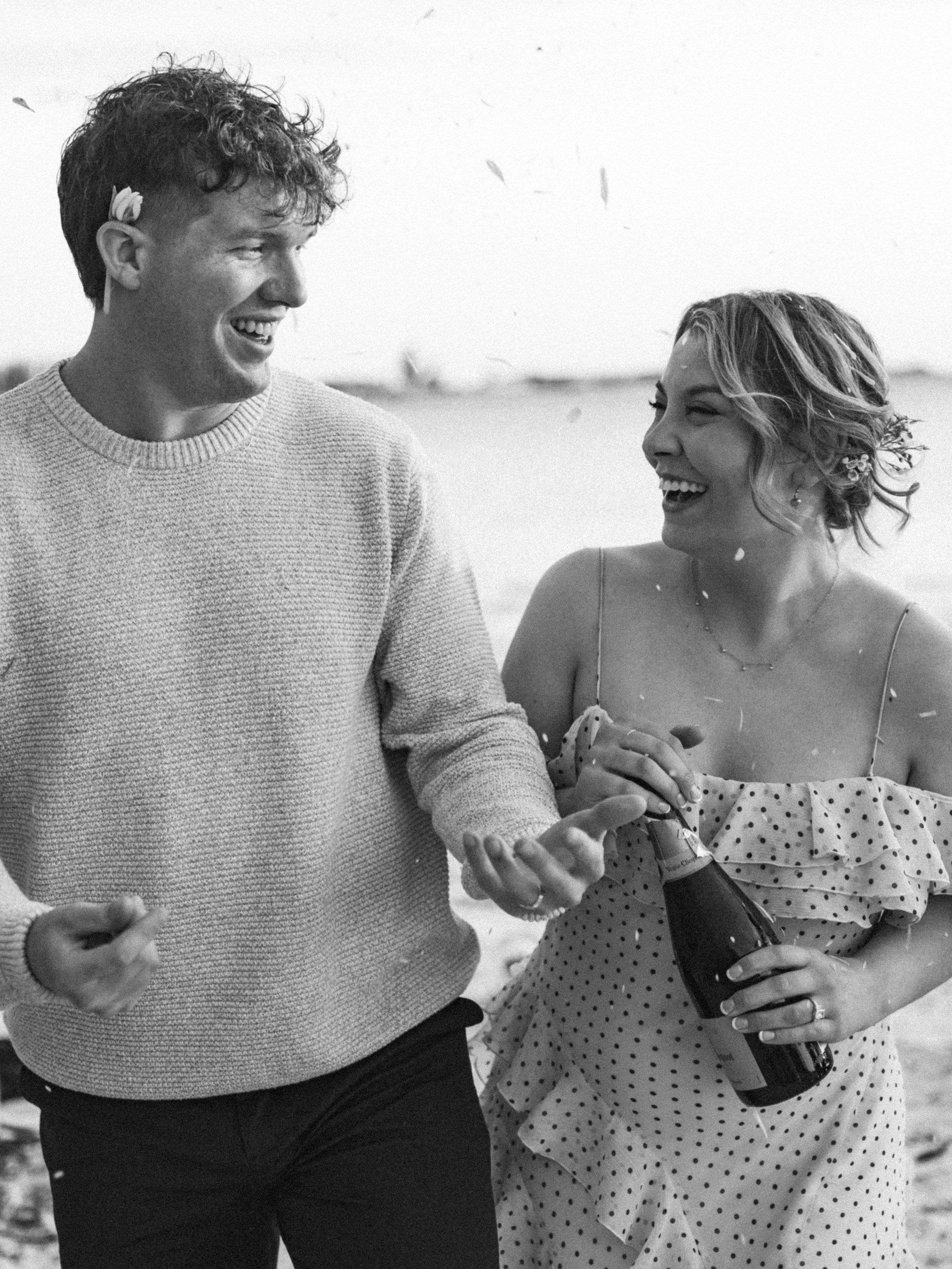 A young man and woman laughing and enjoying each other's company on a beach. The woman is holding a bottle and they are sharing a joyful moment together.