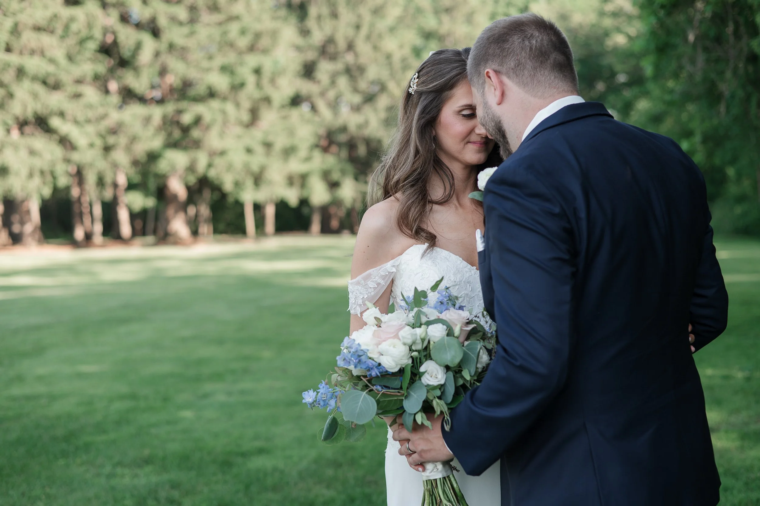 Bride and groom standing close together outdoors, with the bride holding a bouquet of flowers, surrounded by trees and greenery.