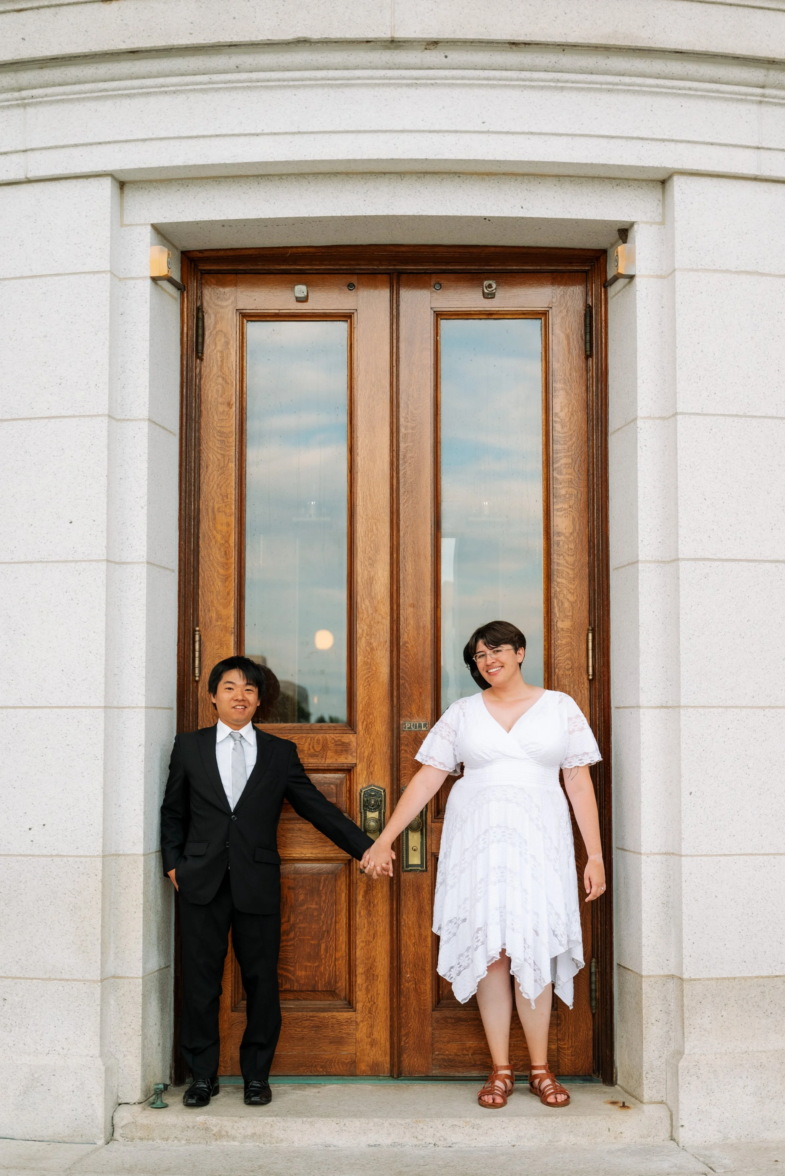 A man in a suit and a woman in a white dress holding hands and smiling while standing in front of a large wooden door.