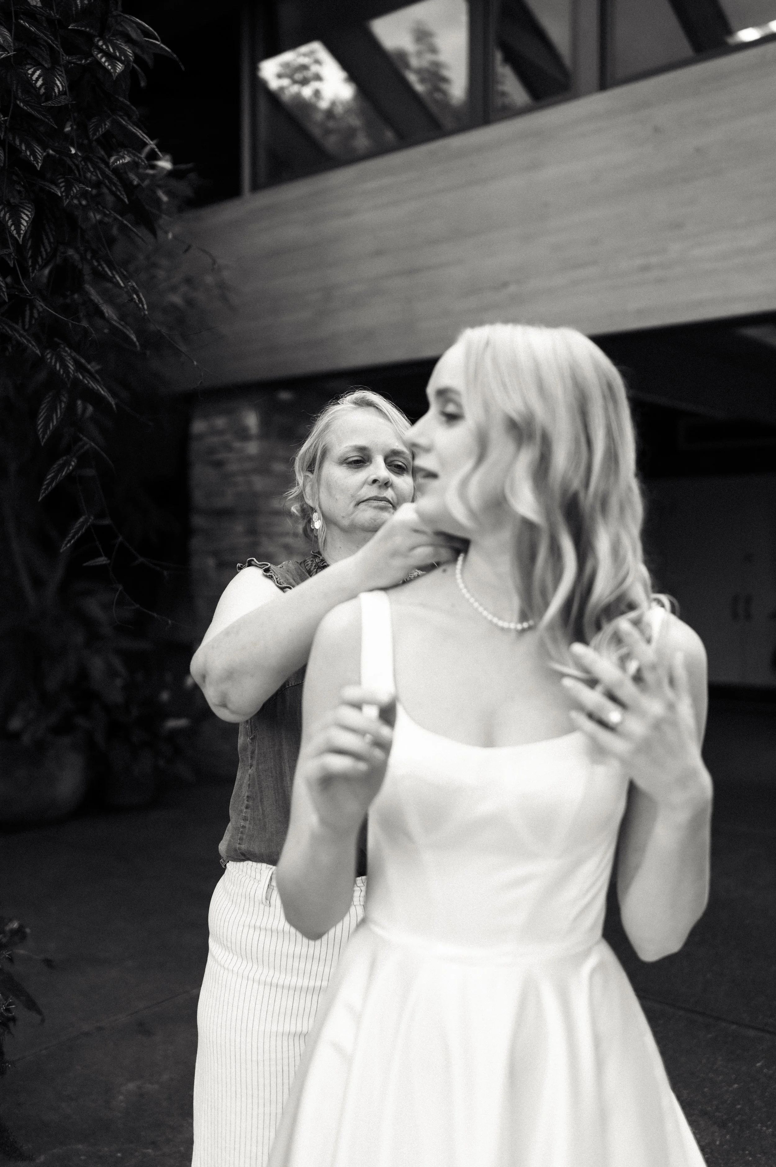 An older woman helps a young woman get ready, adjusting her necklace before she wears a white dress for a special occasion indoors.