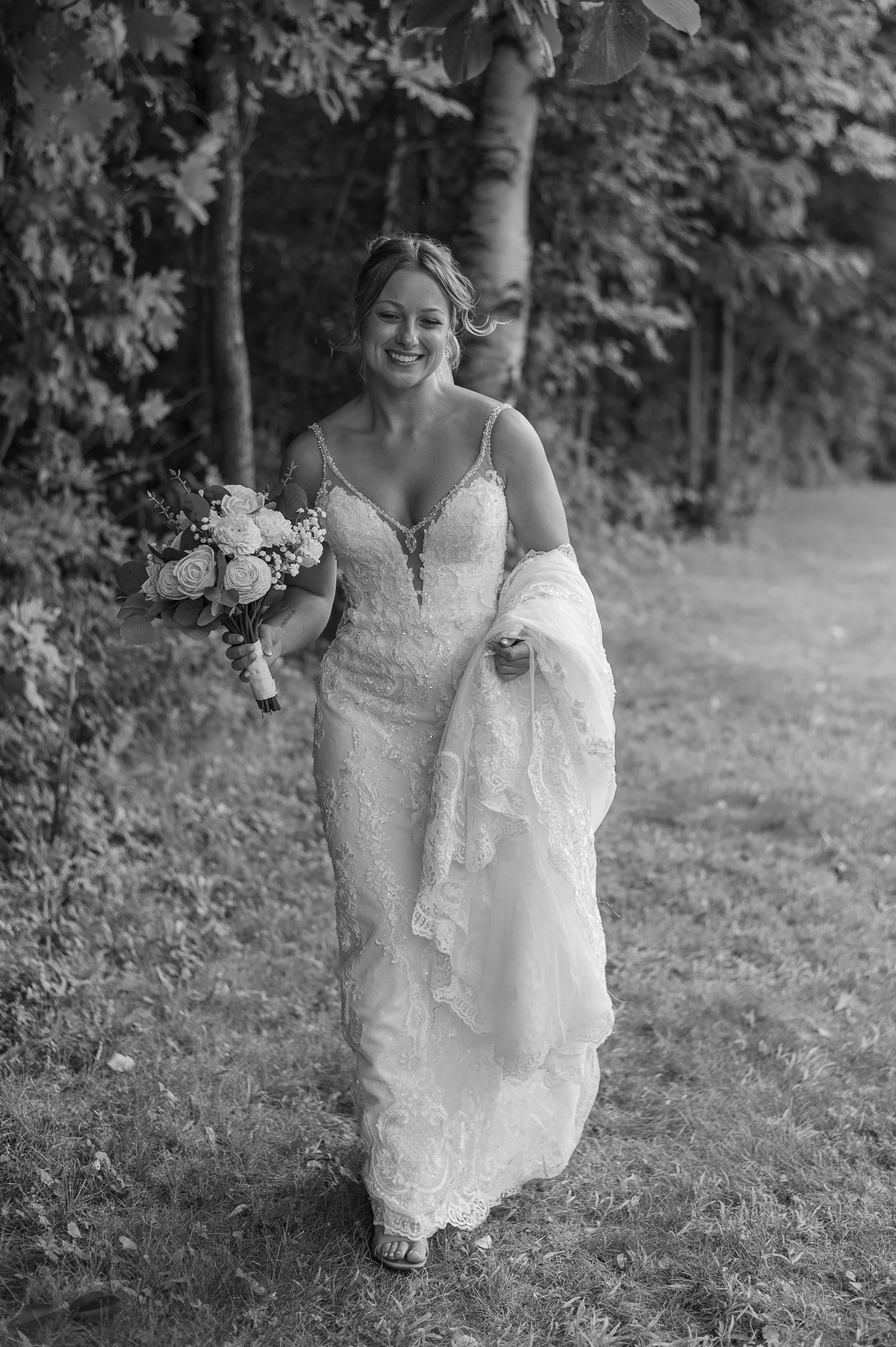 A bride walking outdoors on a grassy path, holding a bouquet of flowers, wearing a lace wedding dress, with trees and foliage in the background.