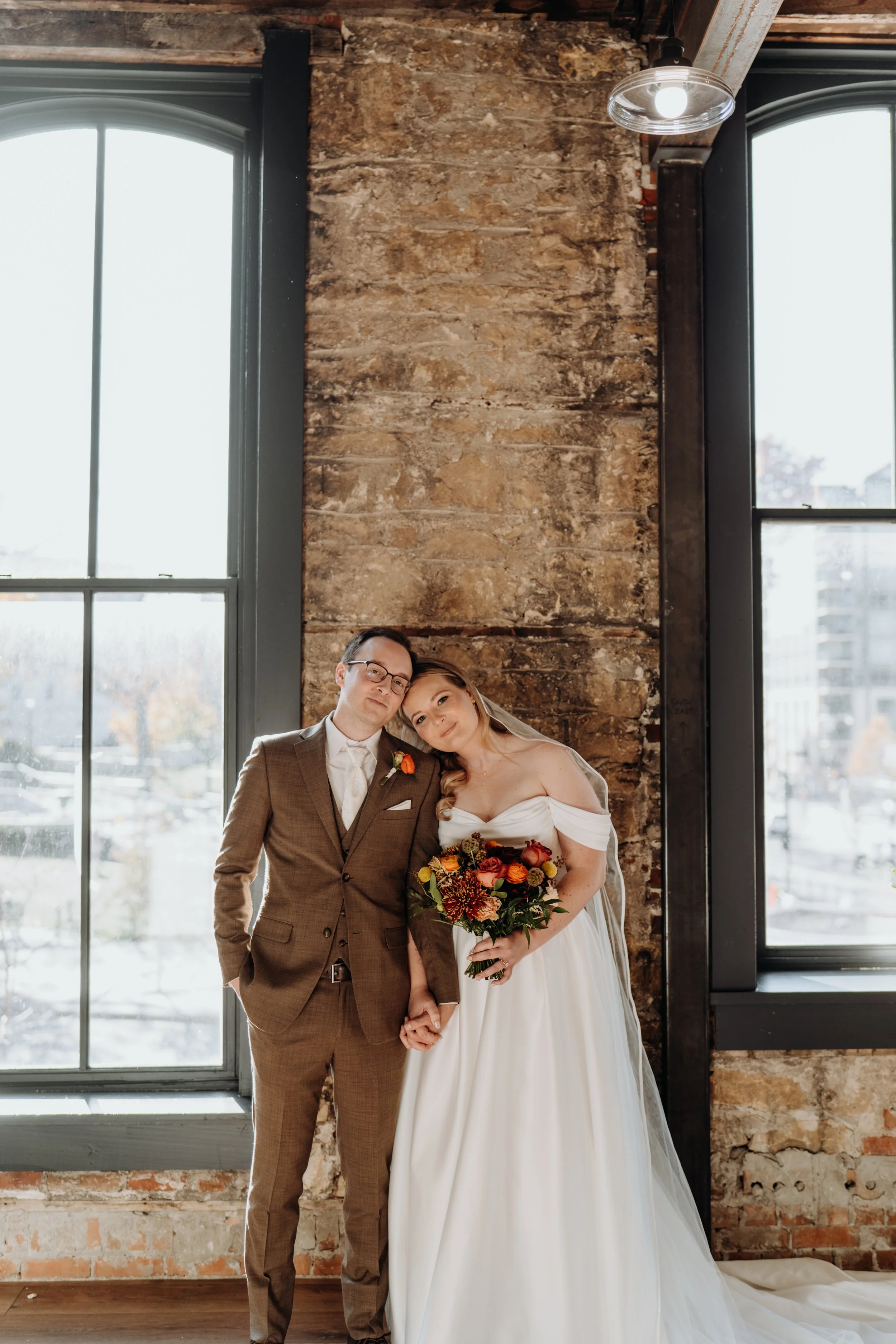 A bride and groom standing together inside a rustic brick building with large black-framed windows, holding hands and smiling at the camera.