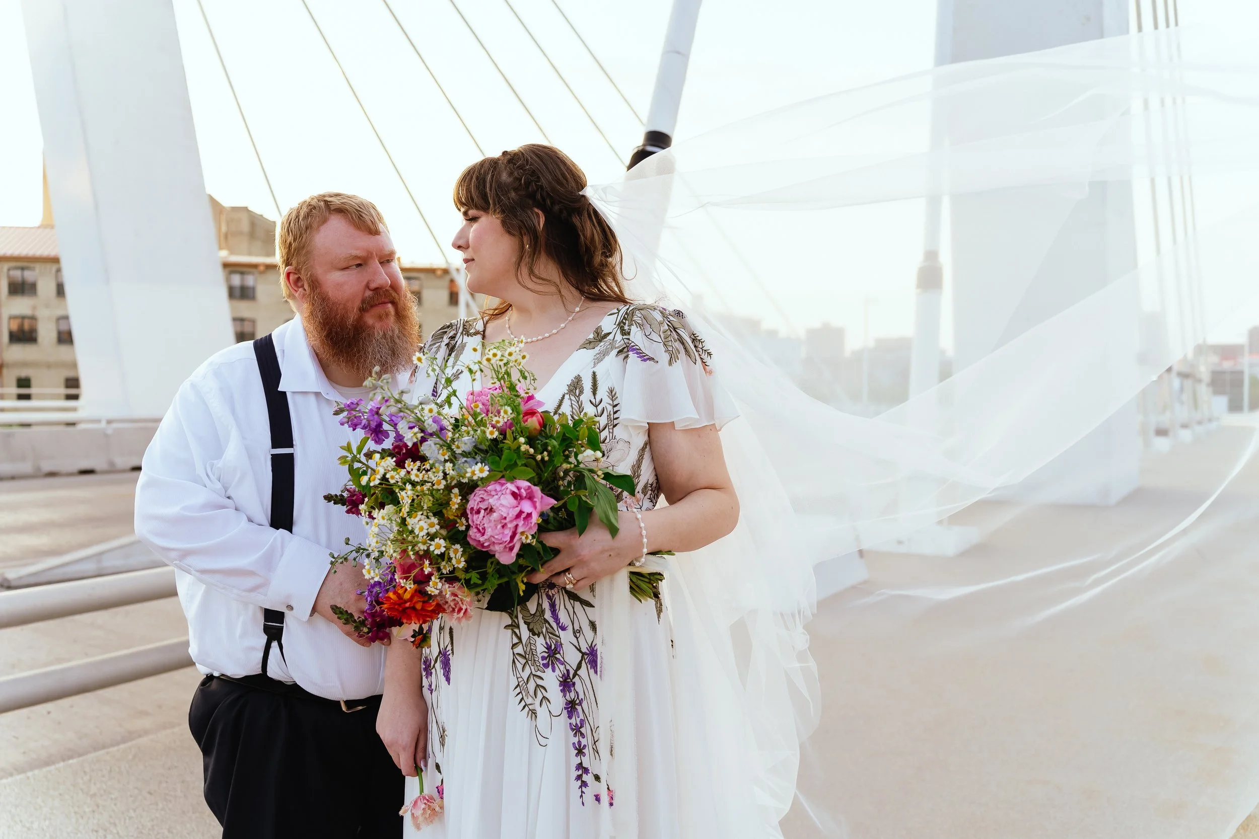 Man with red beard and a woman in a floral dress holding a bouquet of colorful flowers, standing together outdoors near a modern architecturally designed structure.
