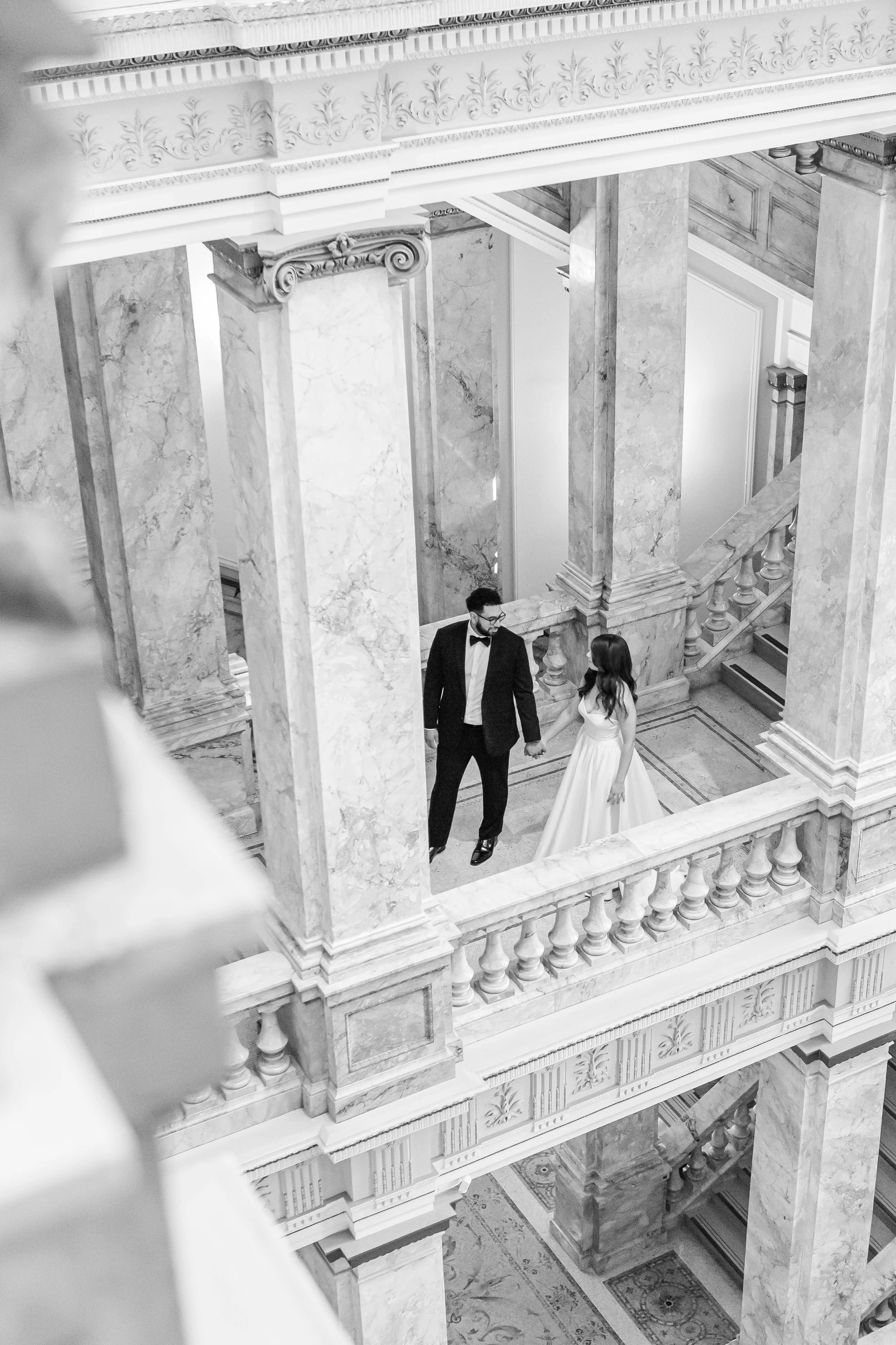A black and white photo of a couple holding hands on a marble balcony in an elegant, historic building, with ornate columns and decorative architectural details.