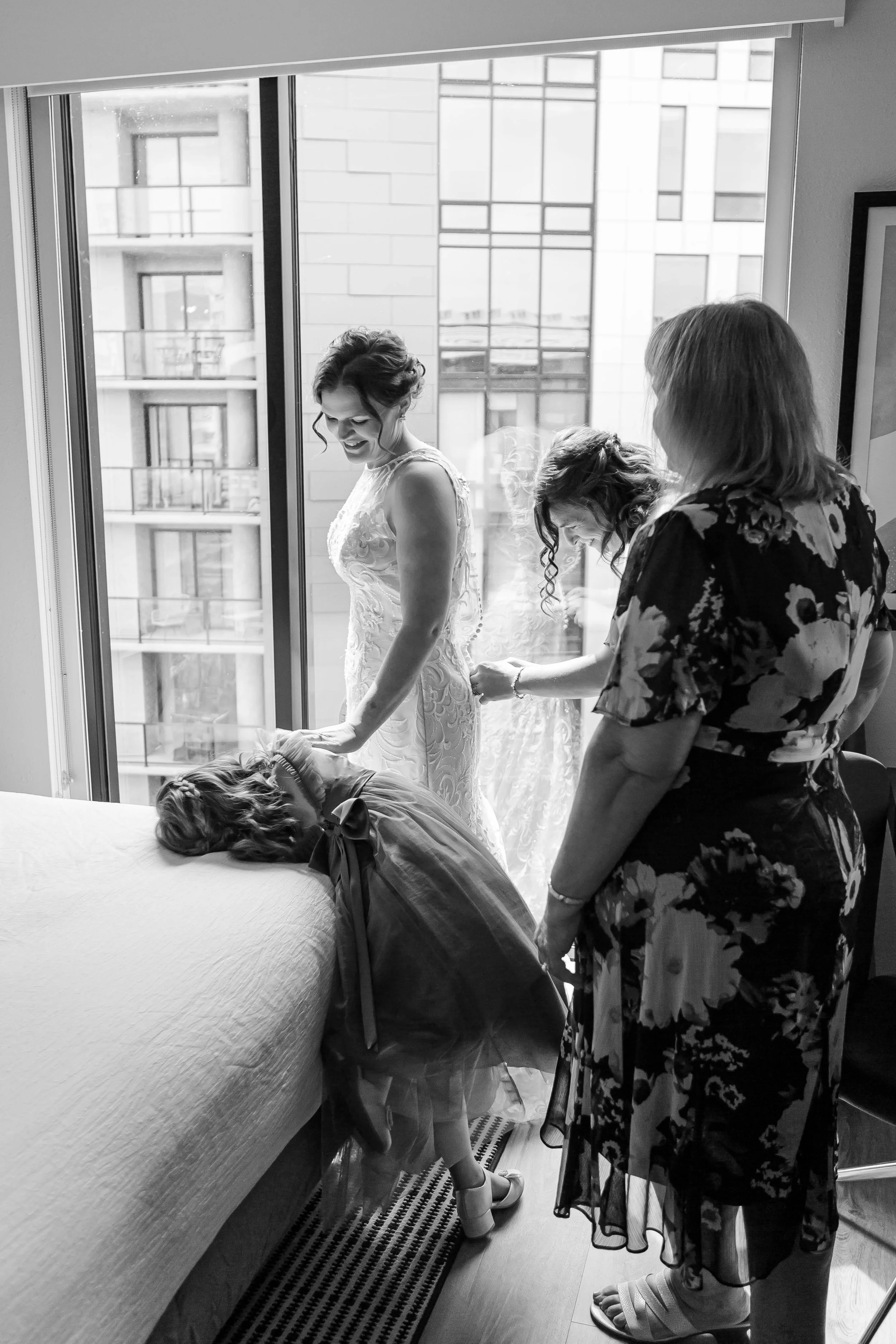 A bride in a wedding dress and a young girl are in a hotel room, with the girl bending over the bed. Two women are helping the bride, with one adjusting her dress. They are near a large window showing a cityscape outside.