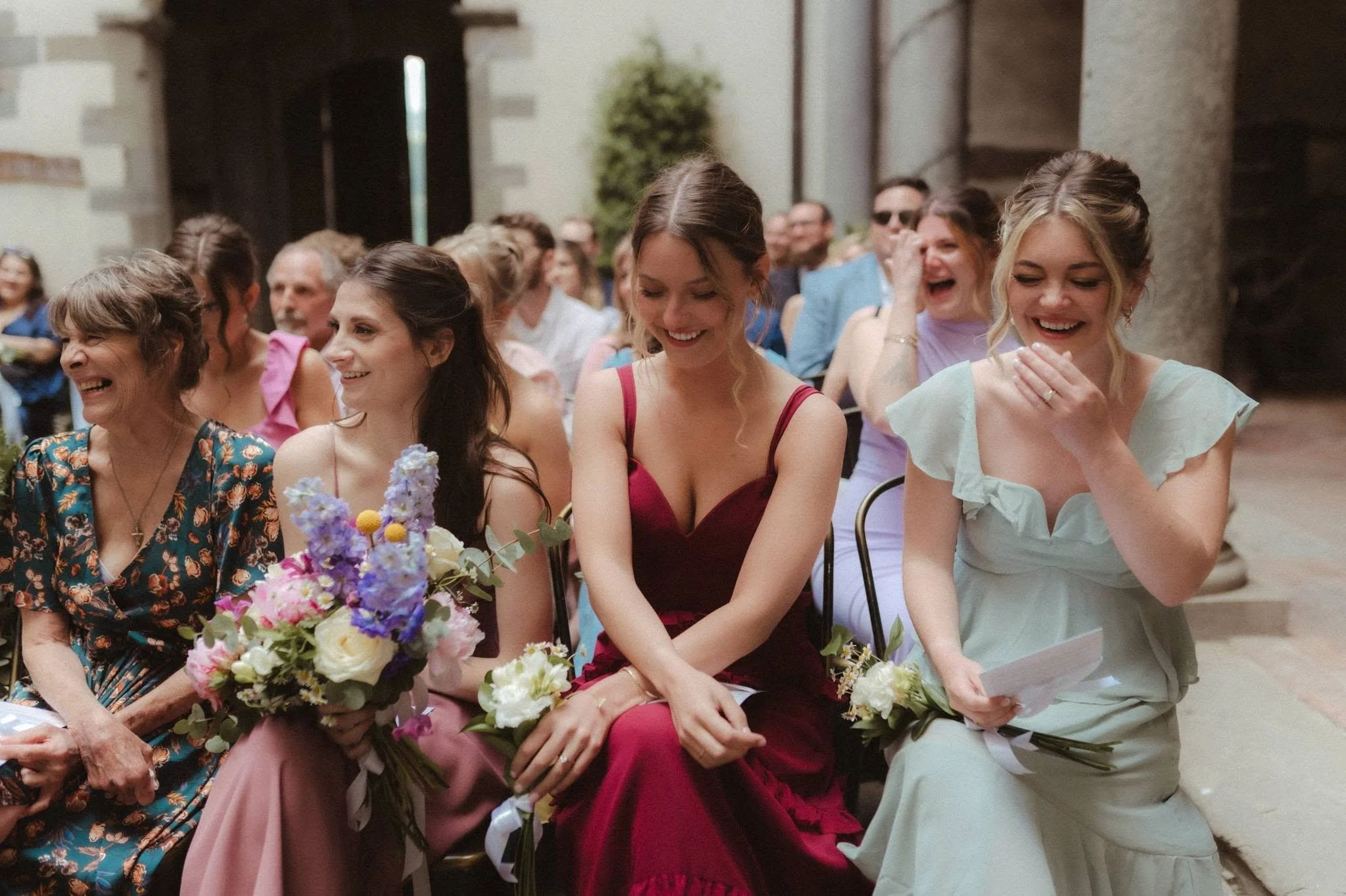 Group of women dressed in formal attire sitting with bouquets of flowers, laughing and smiling at an indoor event.
