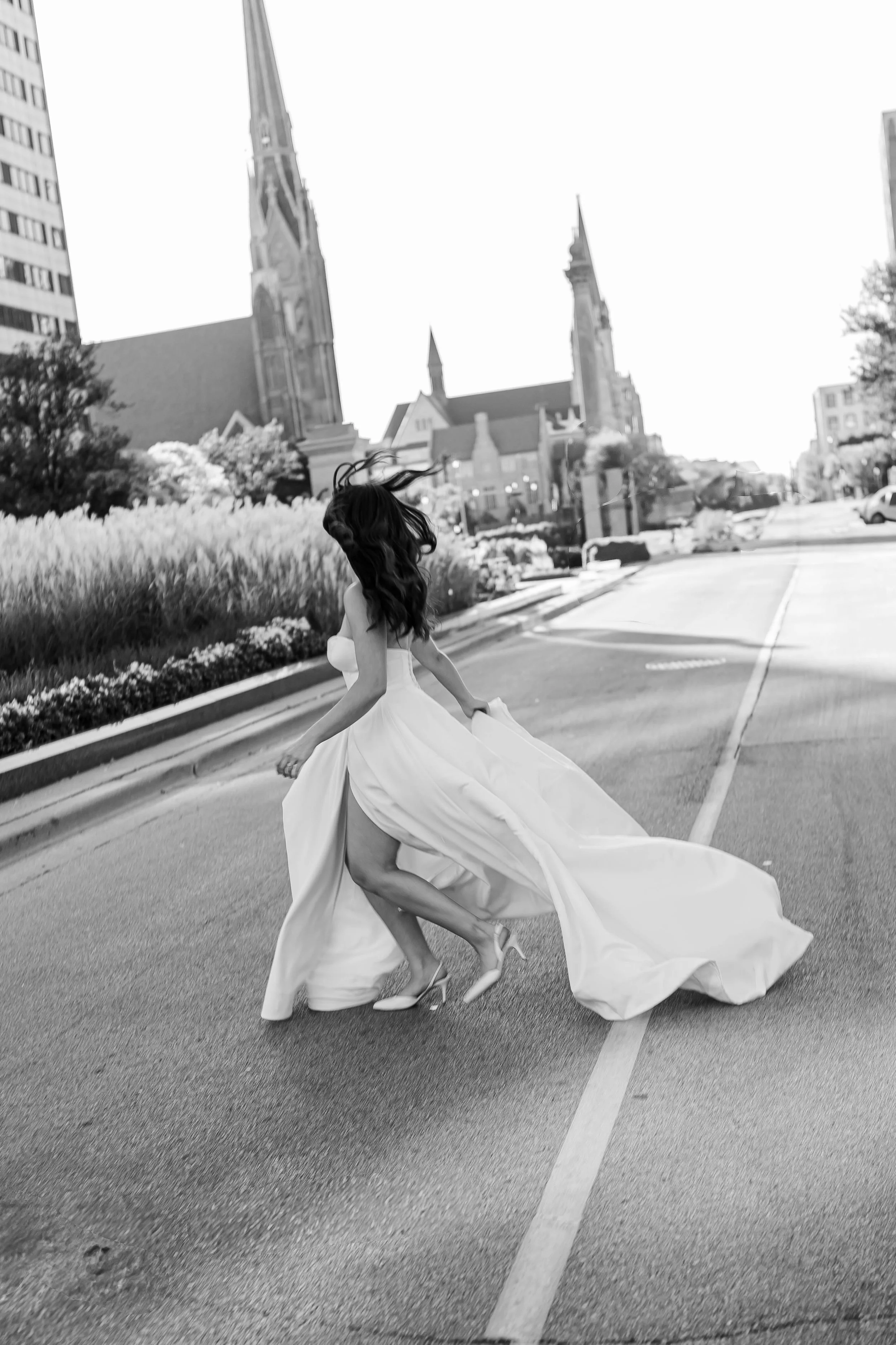 A woman in a wedding dress dancing on an empty city street, with a church and tall buildings in the background.