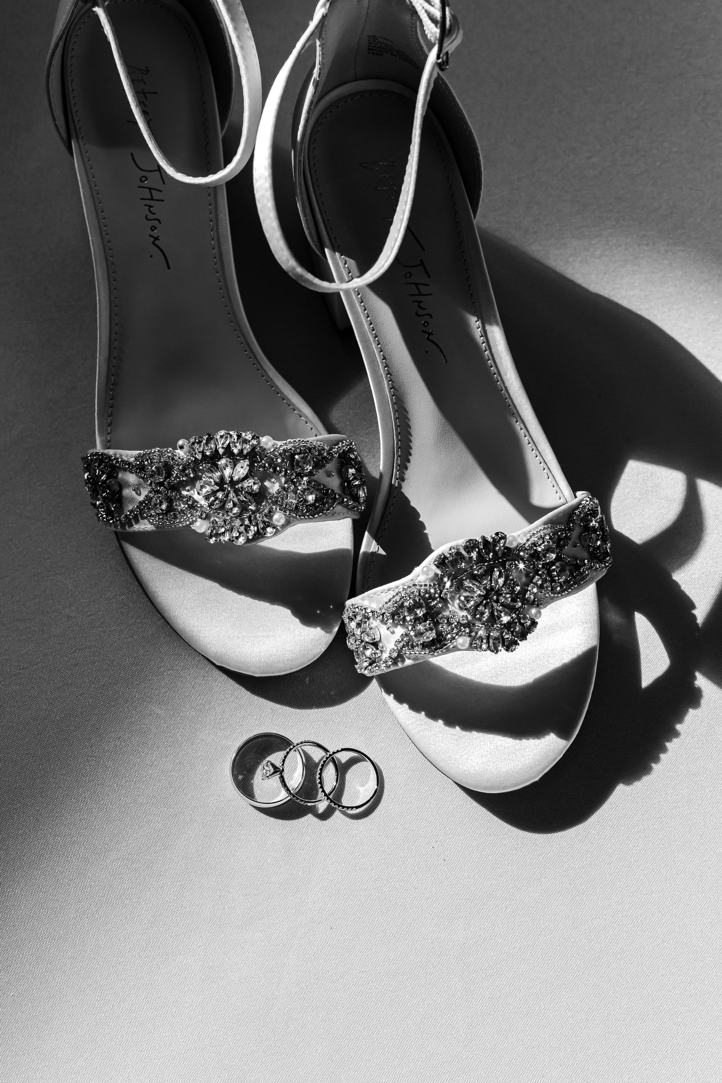 A pair of elegant high-heeled shoes with jewel-encrusted straps, a wedding ring, an engagement ring, and a simple band ring on a flat surface, with dramatic shadows in a black and white photograph.