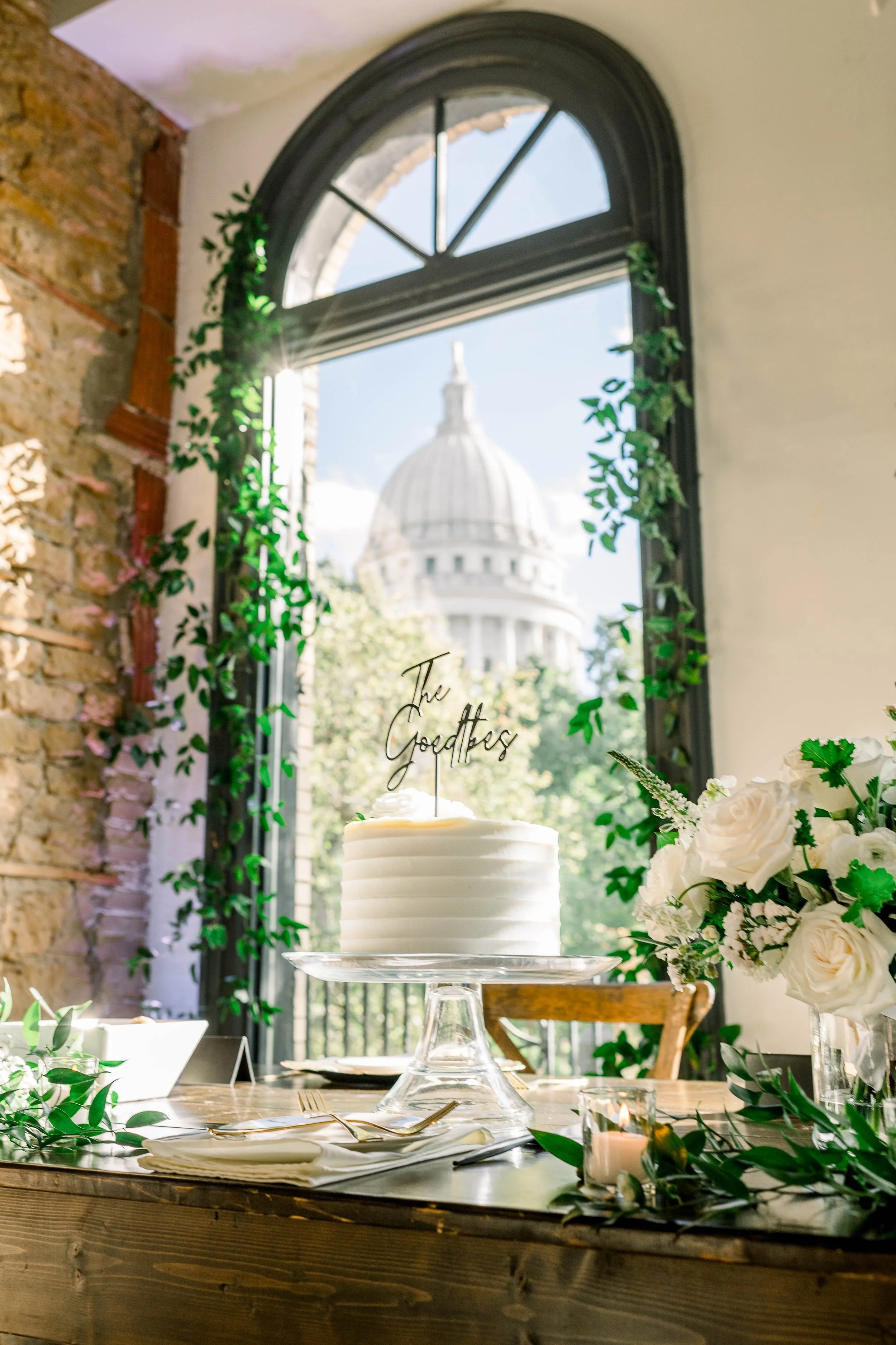 A wedding or celebration cake on a glass cake stand, surrounded by white flowers and green foliage, with a large window showing a domed building outside. The cake topper says "The Goddads."