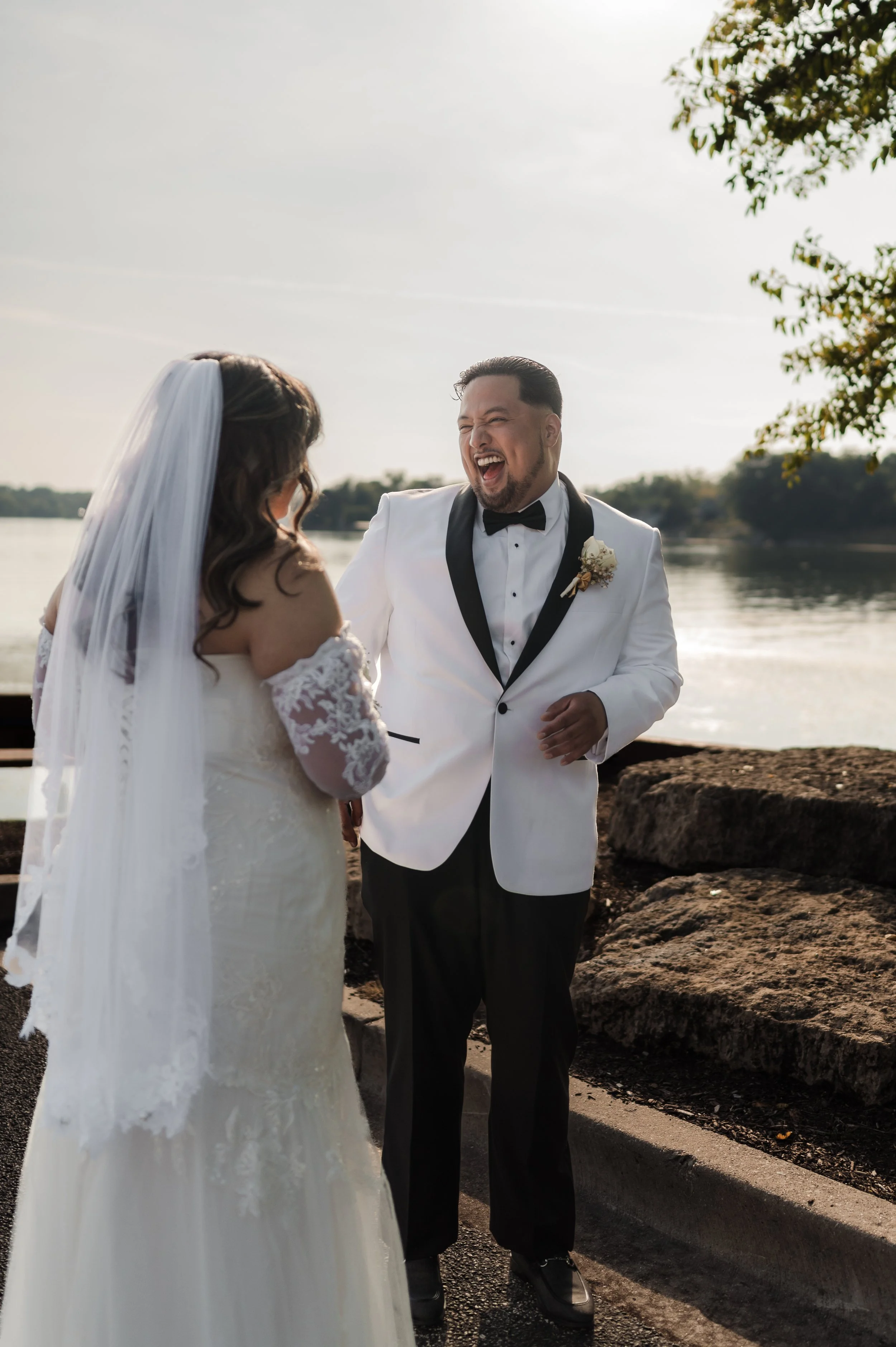 A bride and groom at a wedding ceremony outdoors by a river, sharing a joyful moment with the groom laughing and the bride facing away.