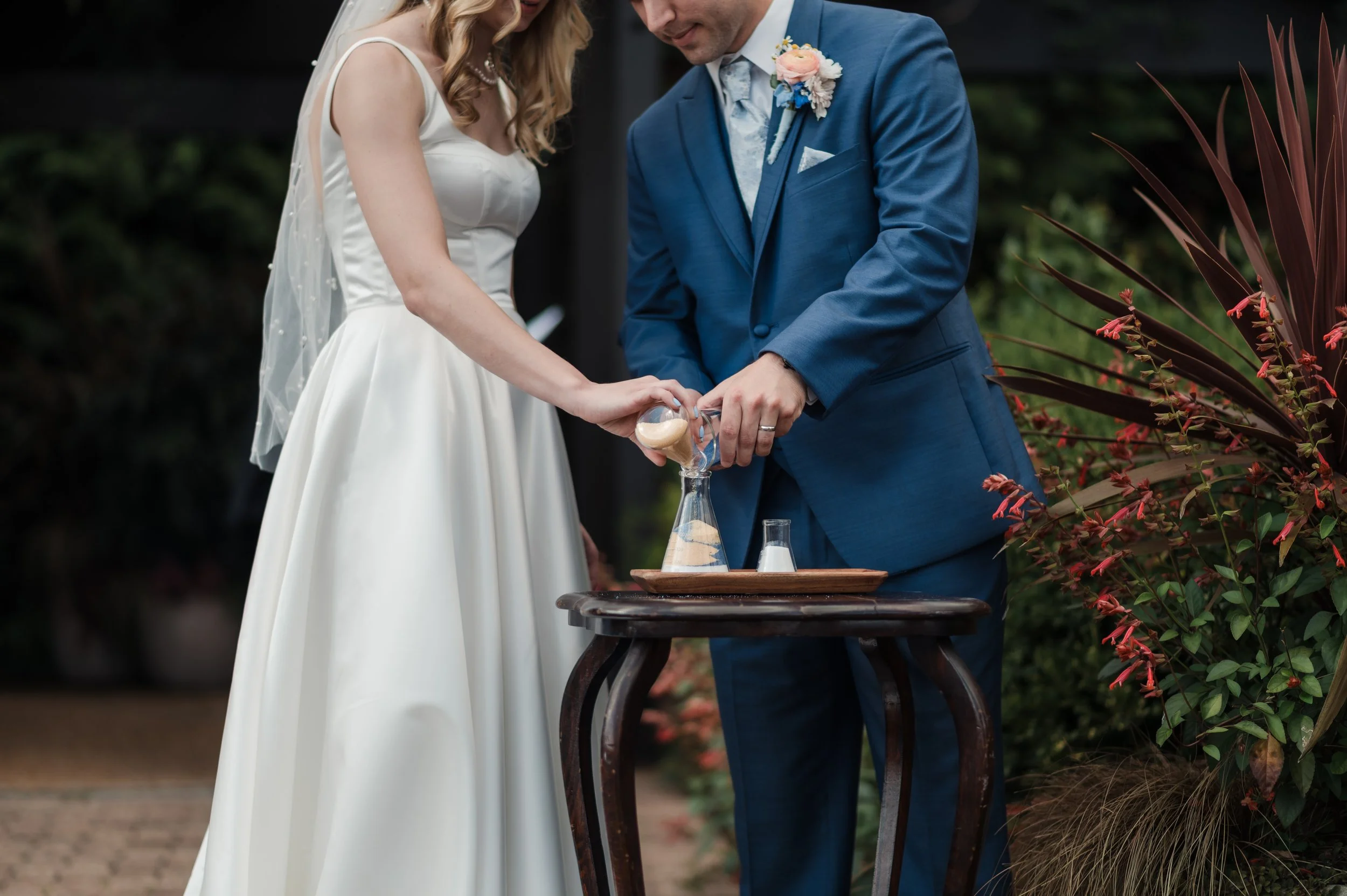 A bride and groom pour sand into a vase during a wedding ceremony outside, with wedding attire, flowers, and greenery.
