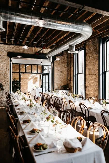 Long banquet table set with white tablecloths, plates, glasses, and floral centerpieces in an industrial-style venue with brick walls, large windows, and exposed ductwork.