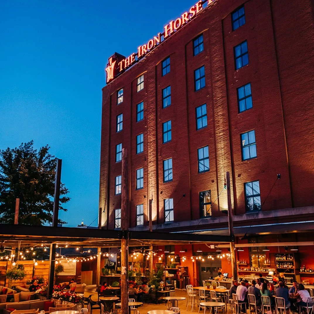 A brick building with a neon sign that says 'The Iron Horse' on top. The building is lit up at night. In front, there is an outdoor seating area with string lights, tables, chairs, and people dining.