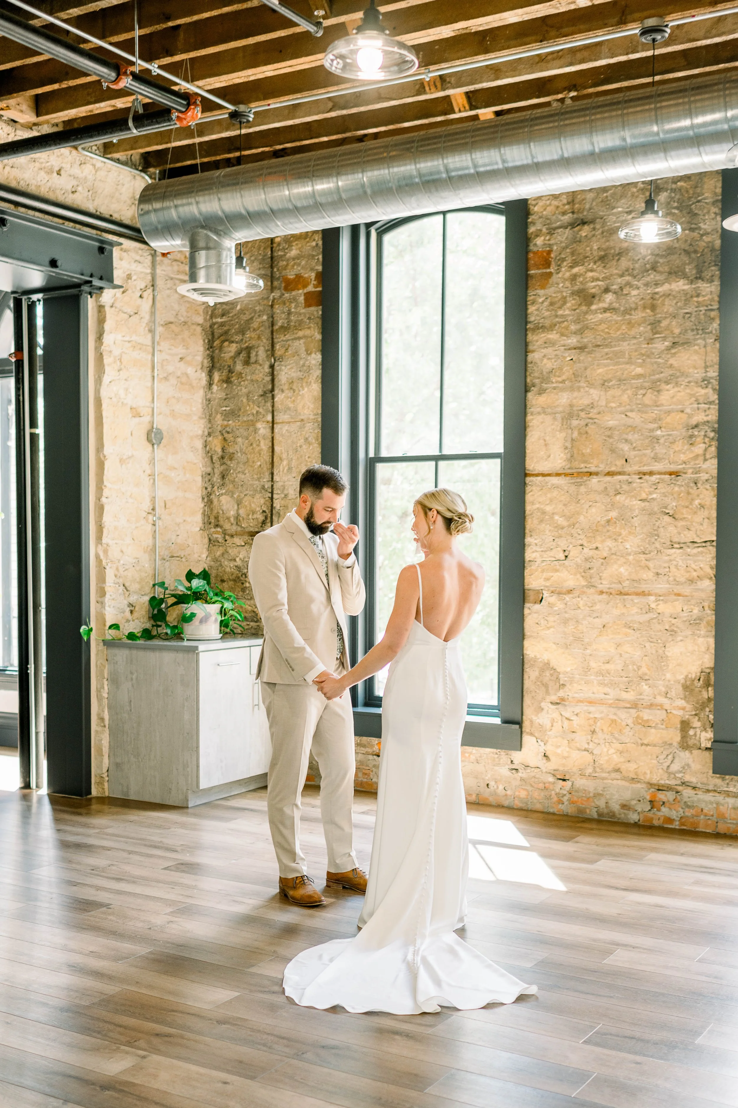 A bride and groom exchange vows in an industrial-style wedding venue with exposed brick walls, large windows, and wooden floors. The bride wears a white gown with a train, and the groom is in a light suit. They hold hands and look at each other as they share a special moment.