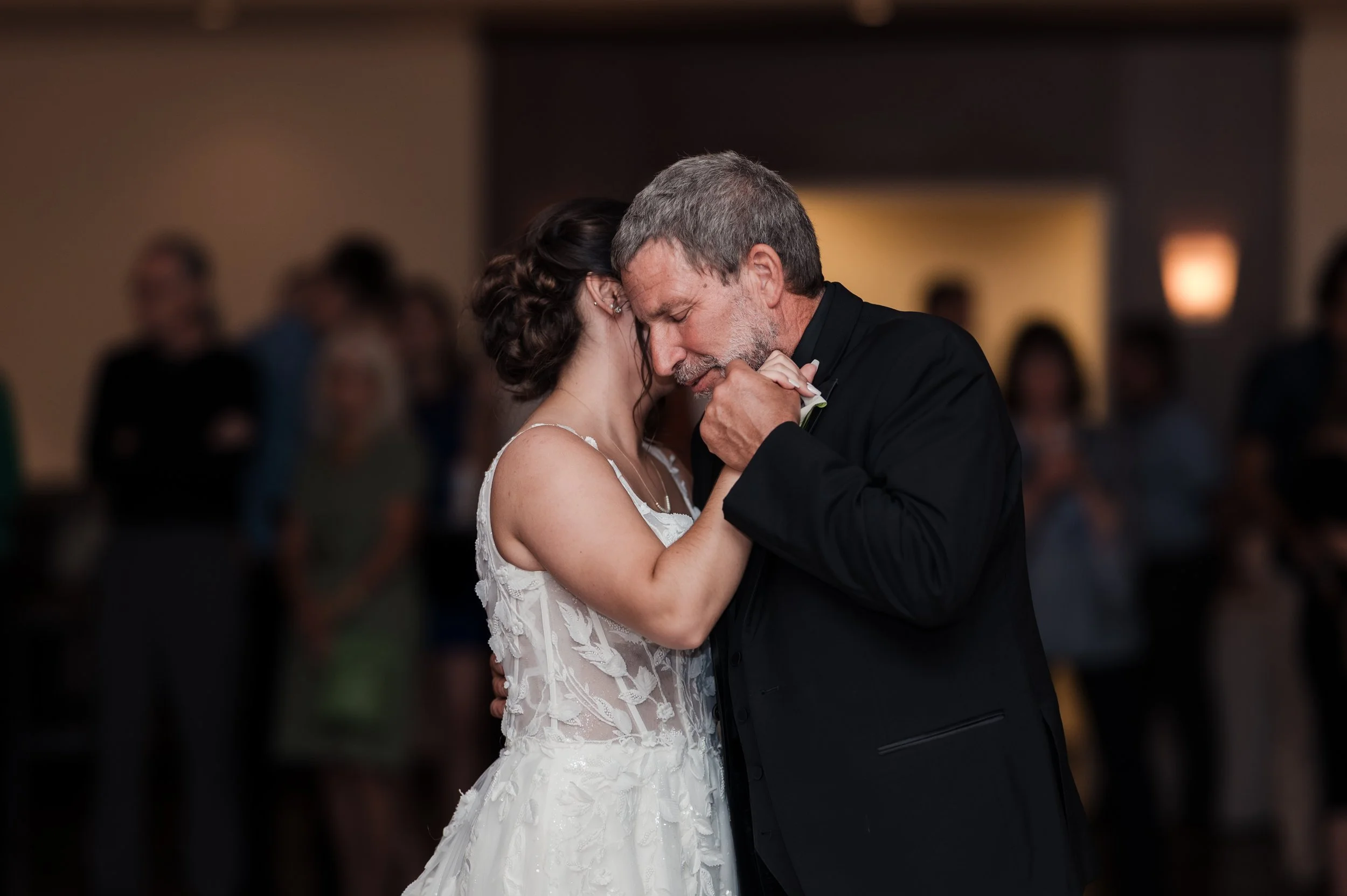 A bride and an older man share a tender dance at a wedding reception, with guests in the background.