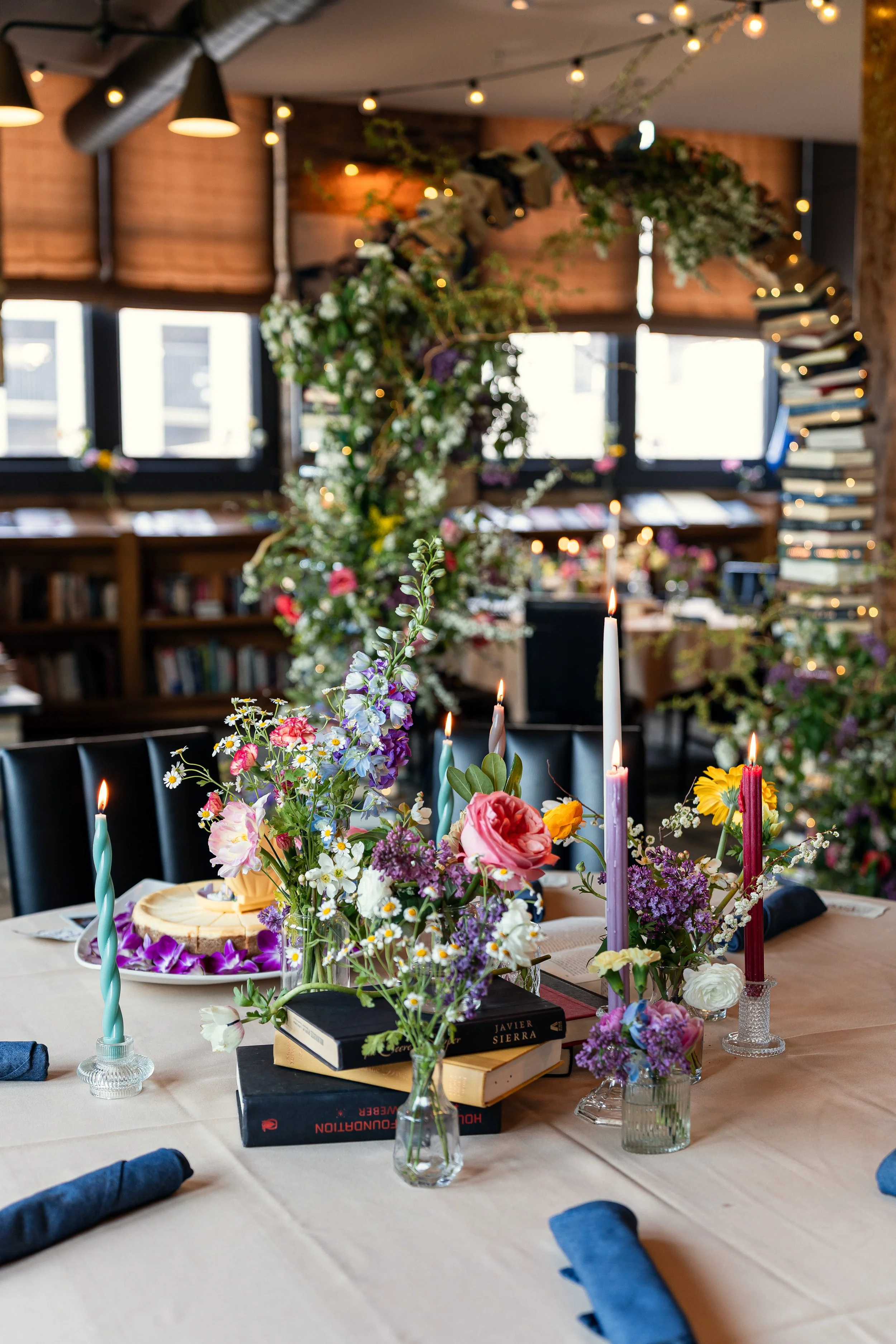 A decorated dining table with floral arrangements, candles, and books, set in a cozy room with large windows, bookshelves, and string lights.