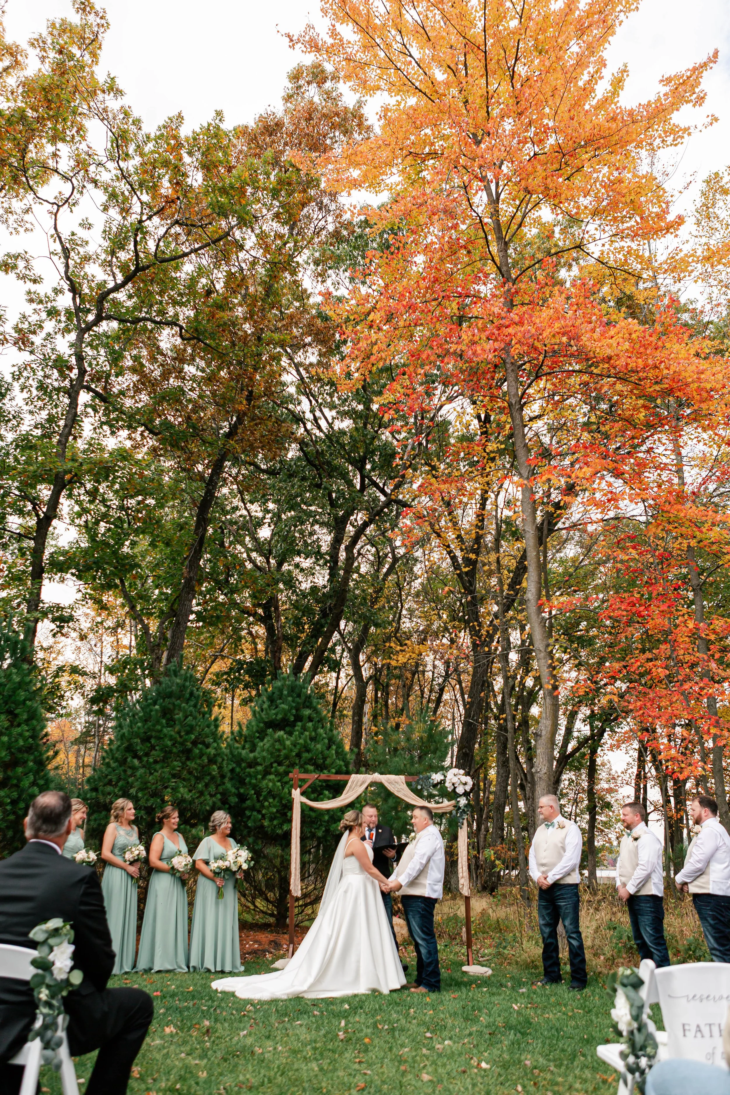A wedding ceremony taking place outdoors in a fall setting, with the bride and groom holding hands under a decorated arch, surrounded by bridesmaids in pastel dresses and groomsmen in vests and shirts, and guests seated on decorated chairs, with colo