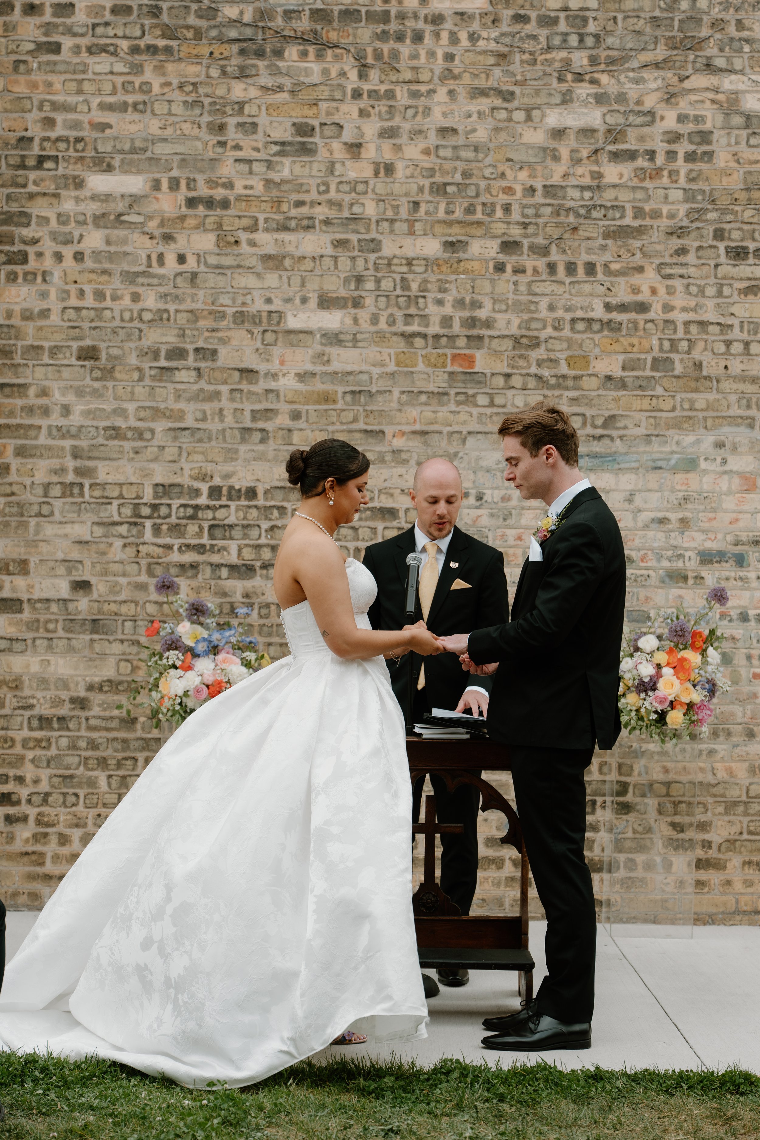 A bride and groom holding hands during their wedding ceremony with a officiant standing between them, against a brick wall backdrop, surrounded by colorful flower arrangements.
