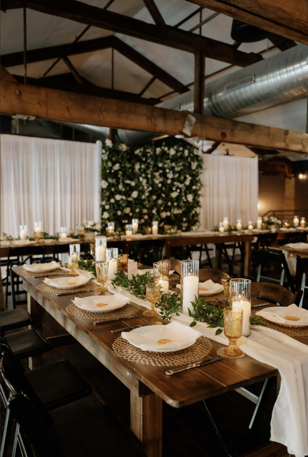 A decorated banquet table set for a formal event in an industrial-style venue with exposed wooden beams and ductwork, featuring candles, greenery, and place settings with white napkins and gold-rimmed glasses.