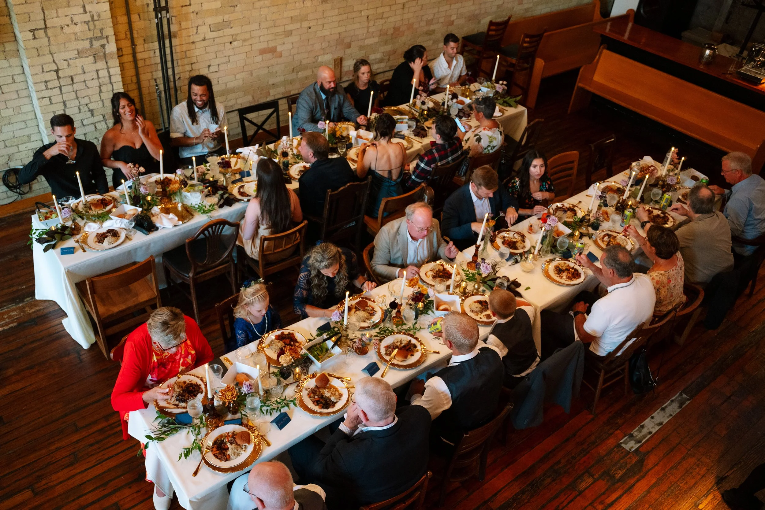 People sitting at long banquet tables decorated with candles, flowers, and plates of food, enjoying a meal in a rustic indoor setting with exposed brick walls.