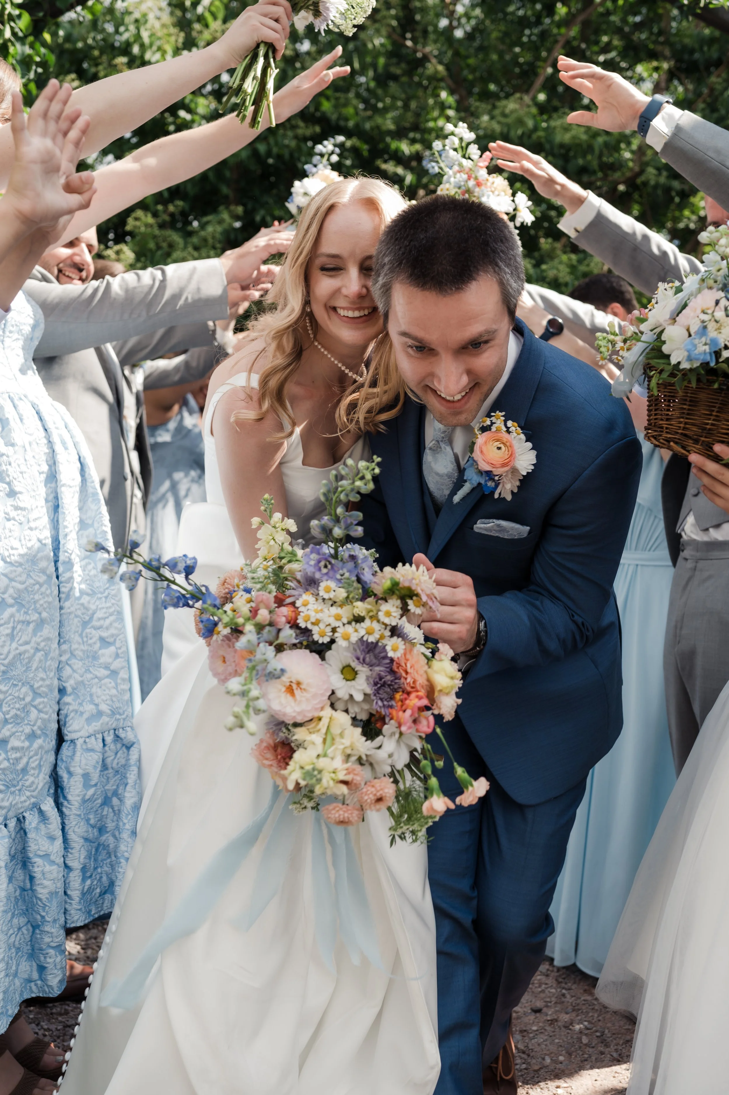 Joyful bride and groom surrounded by friends during their wedding celebration outside, with flowers and greenery.