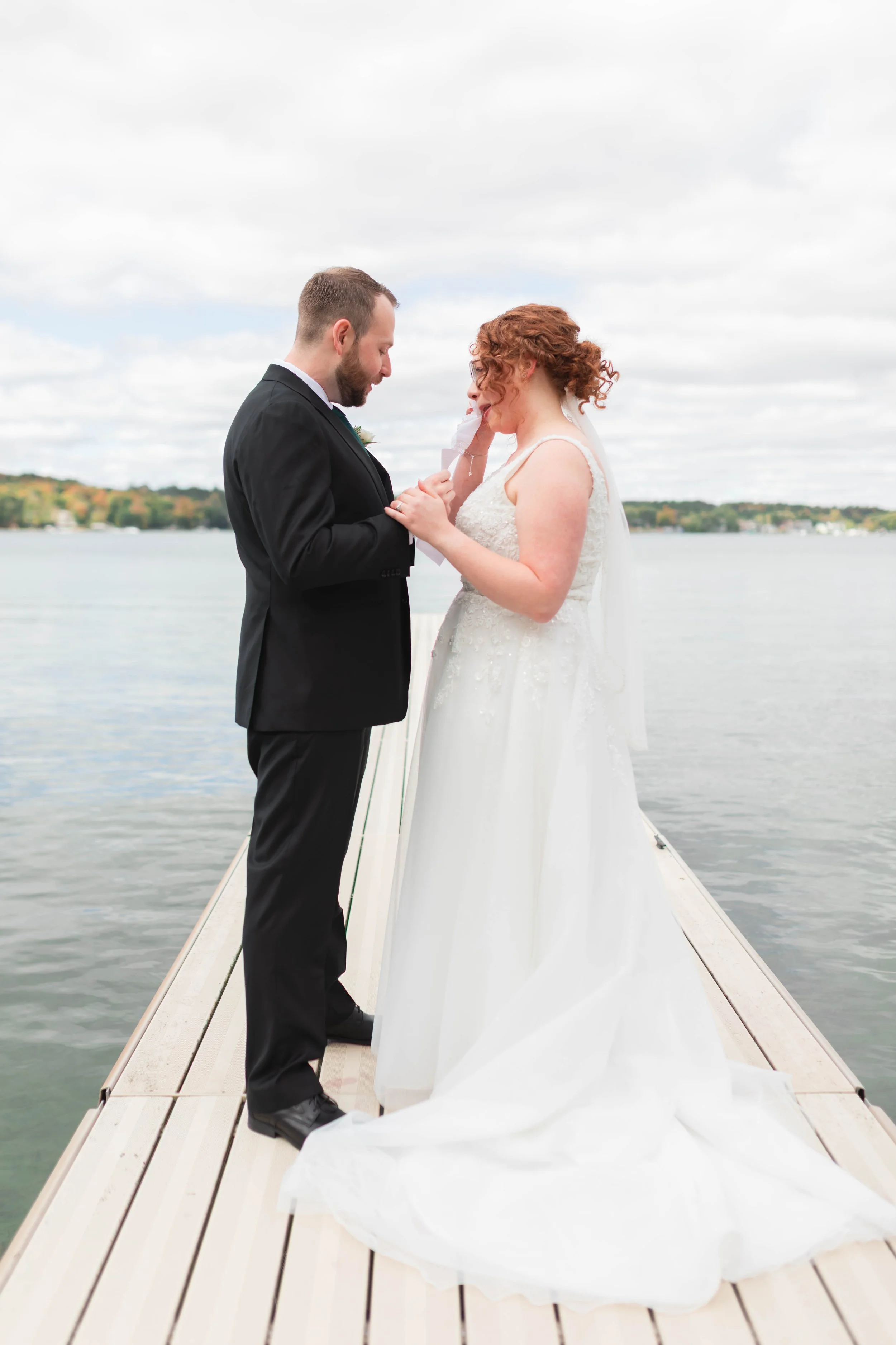 A bride and groom sharing a moment on a dock by a lake, with the bride wiping a tear from her eye as the groom looks at her.