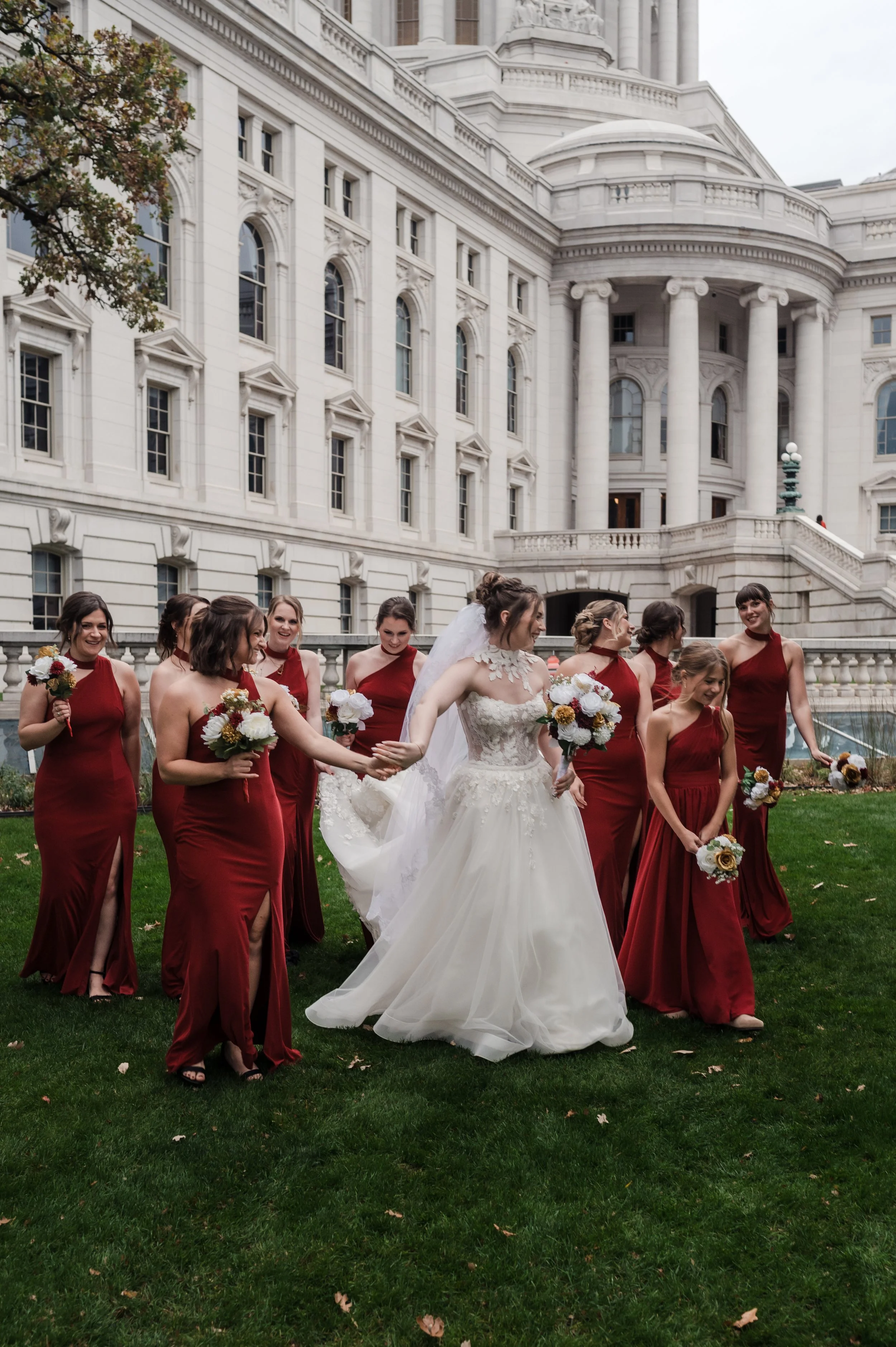 Bride in white wedding dress holding hands with bridesmaids in red dresses, standing on grass outside a large white building with classical architecture.