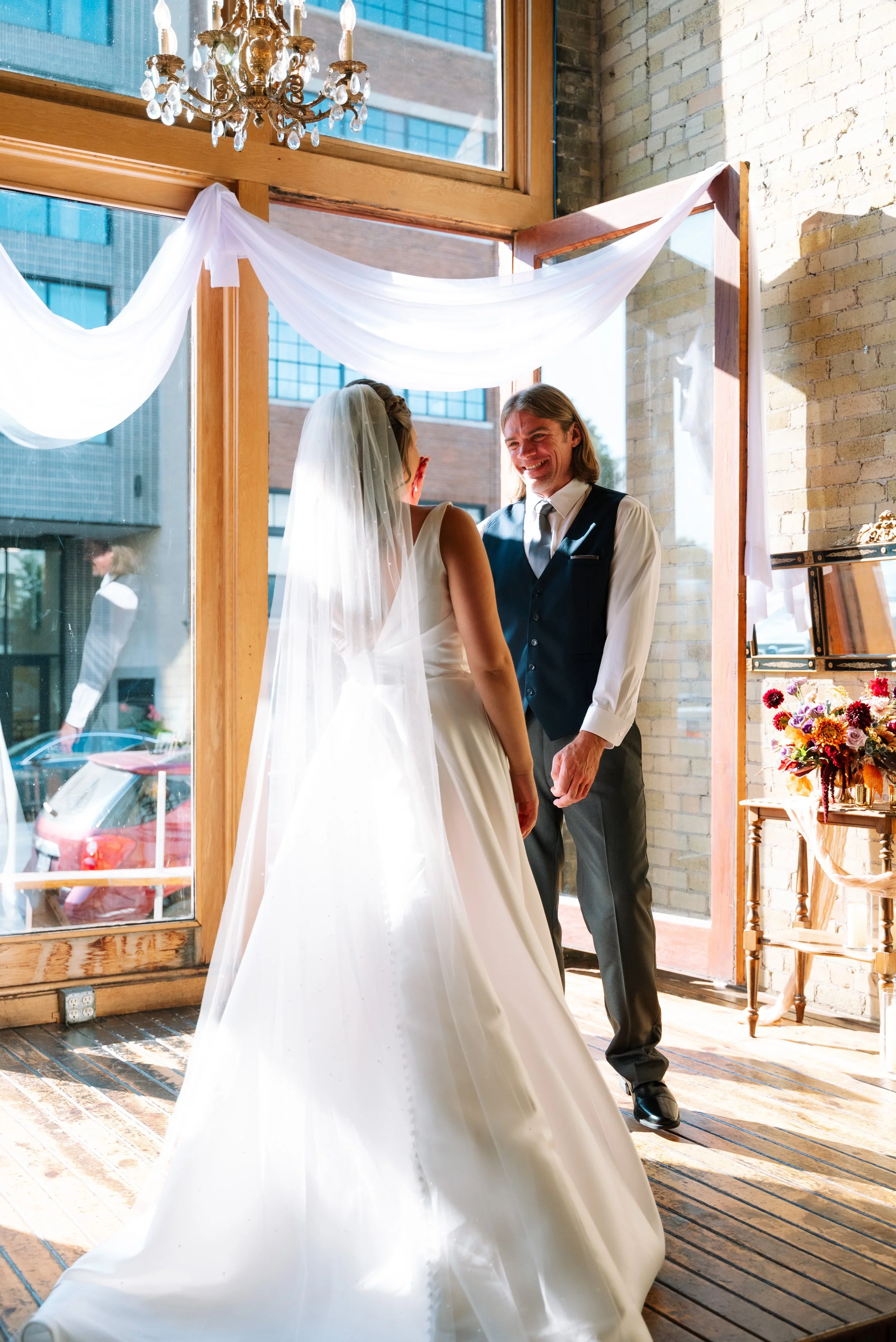 A bride and groom smiling at each other during their wedding ceremony inside a sunny room with large windows, brick walls, wooden floors, and a chandelier. The bride is wearing a white dress and veil, and the groom is in a dark vest and dress shirt.