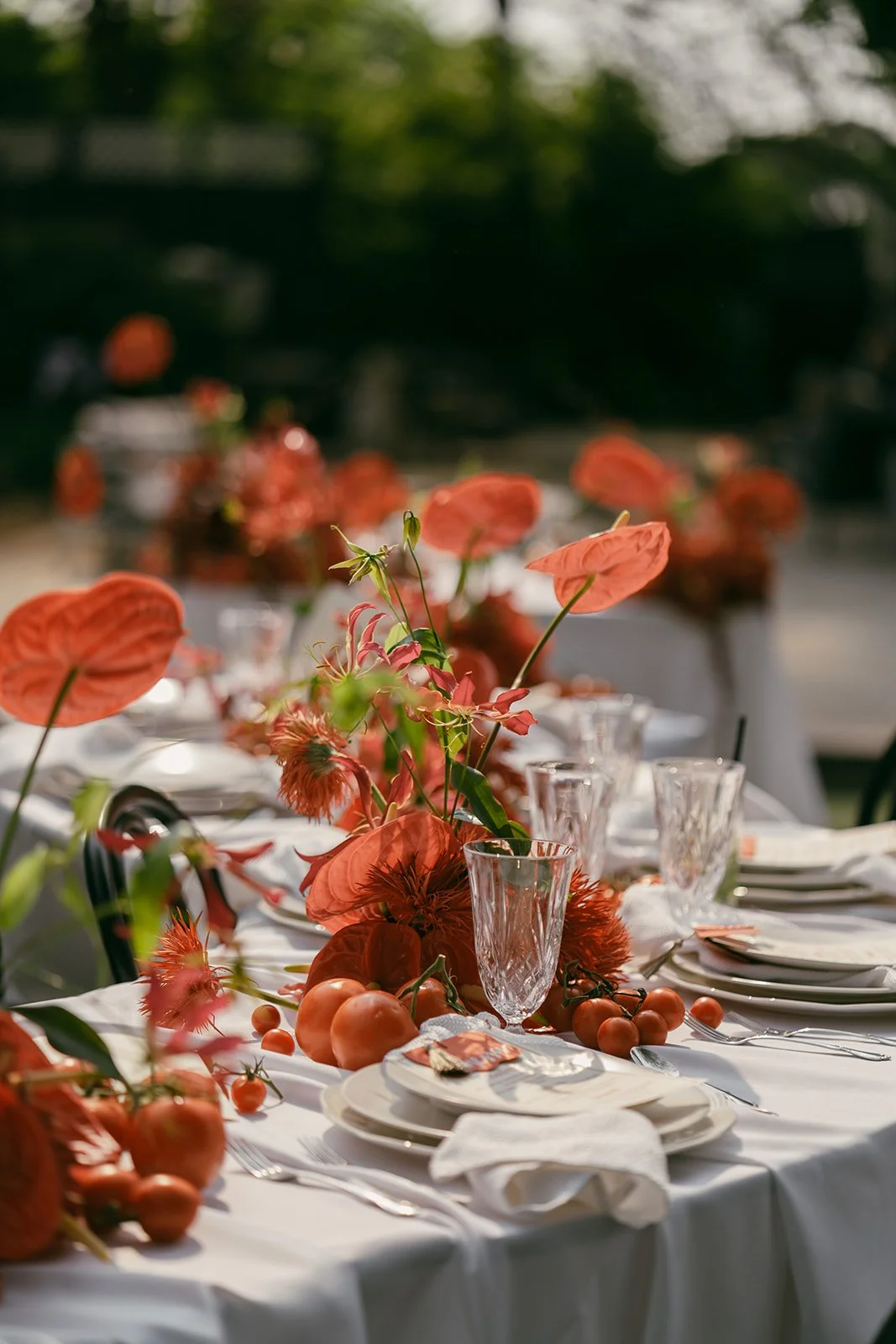 Elegant outdoor dining table decorated with orange flowers, tomatoes, and glassware, set for a meal in natural sunlight.
