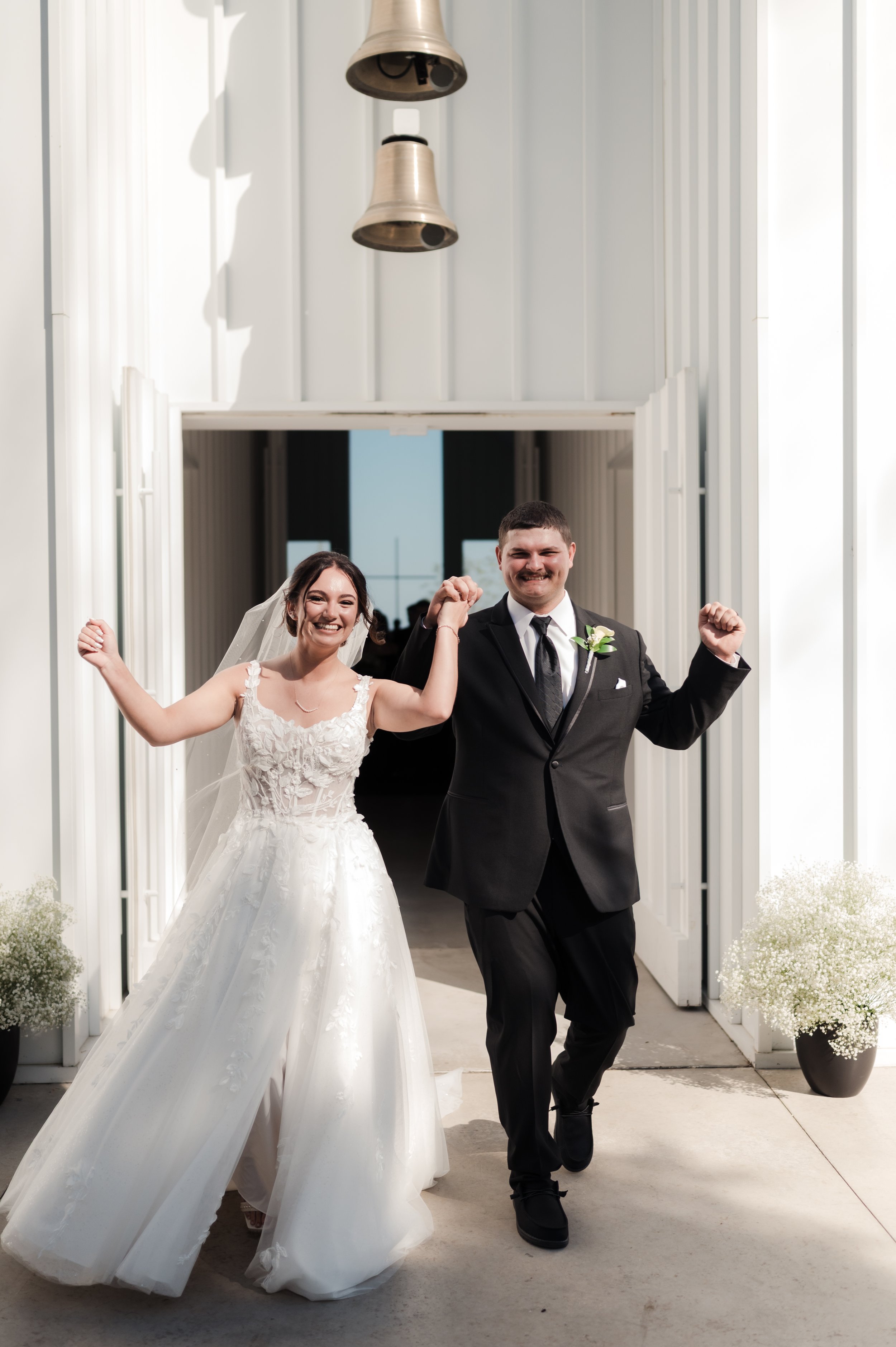 A newlywed couple celebrates as they walk out of a building with their hands raised. The bride is in a white wedding gown with a veil, and the groom is in a black suit with a boutonniere. They are smiling and appear happy.
