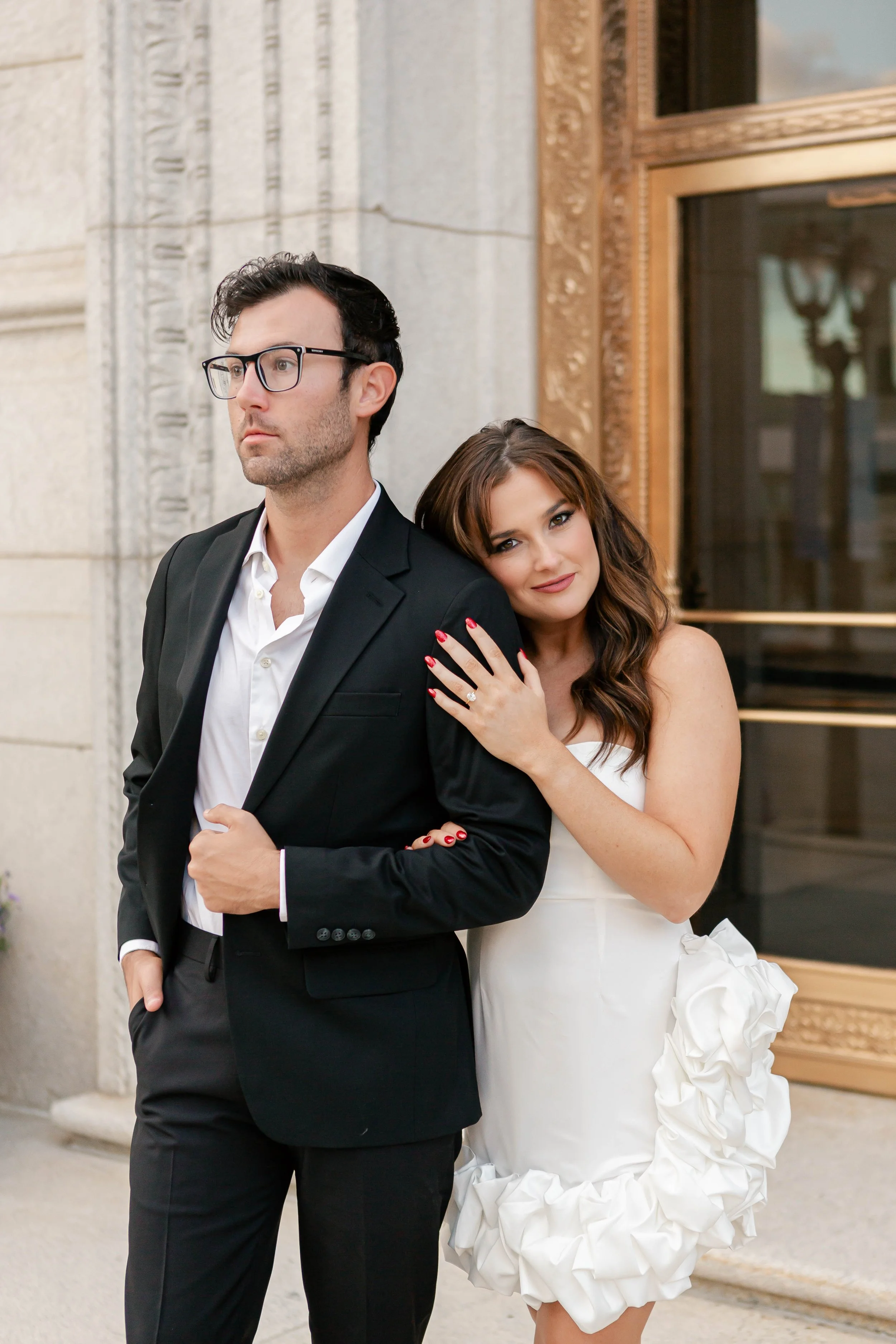 A man and a woman standing outside a building. The woman is leaning her head on the man's shoulder and has her hand on his arm. She is wearing a white dress with ruffles and has long, wavy brown hair. The man is wearing a black suit with a white shir