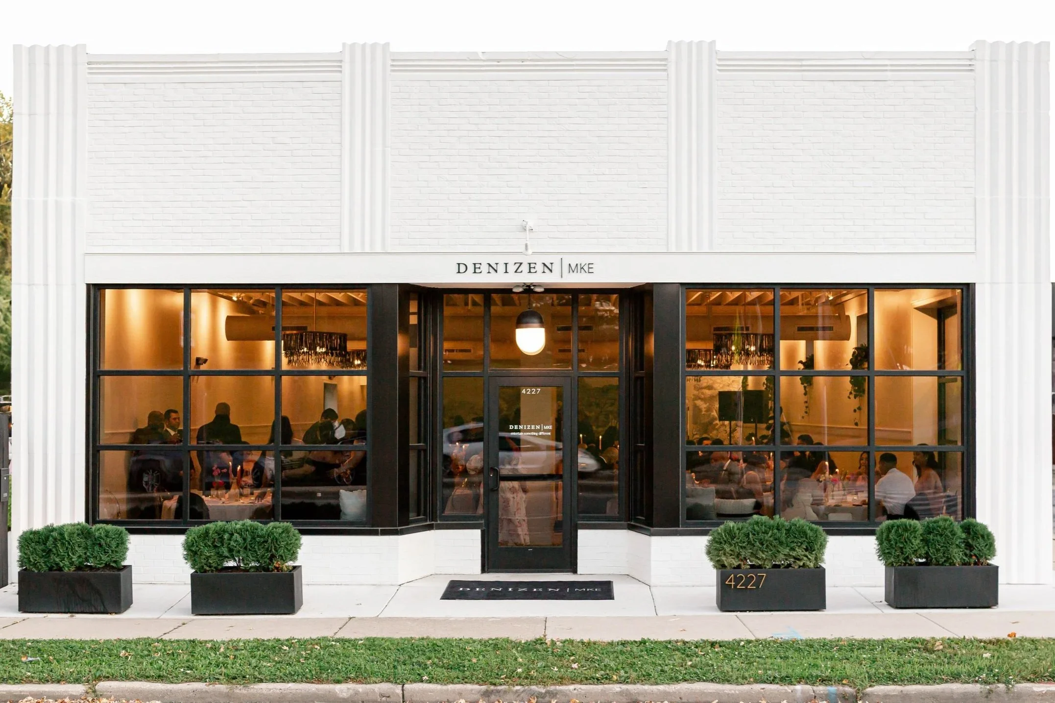 Modern storefront with large glass windows showing a busy interior, white brick exterior, black accents, potted plants outside, and a sign with the name 'DENIZEN MKE' above the entrance.