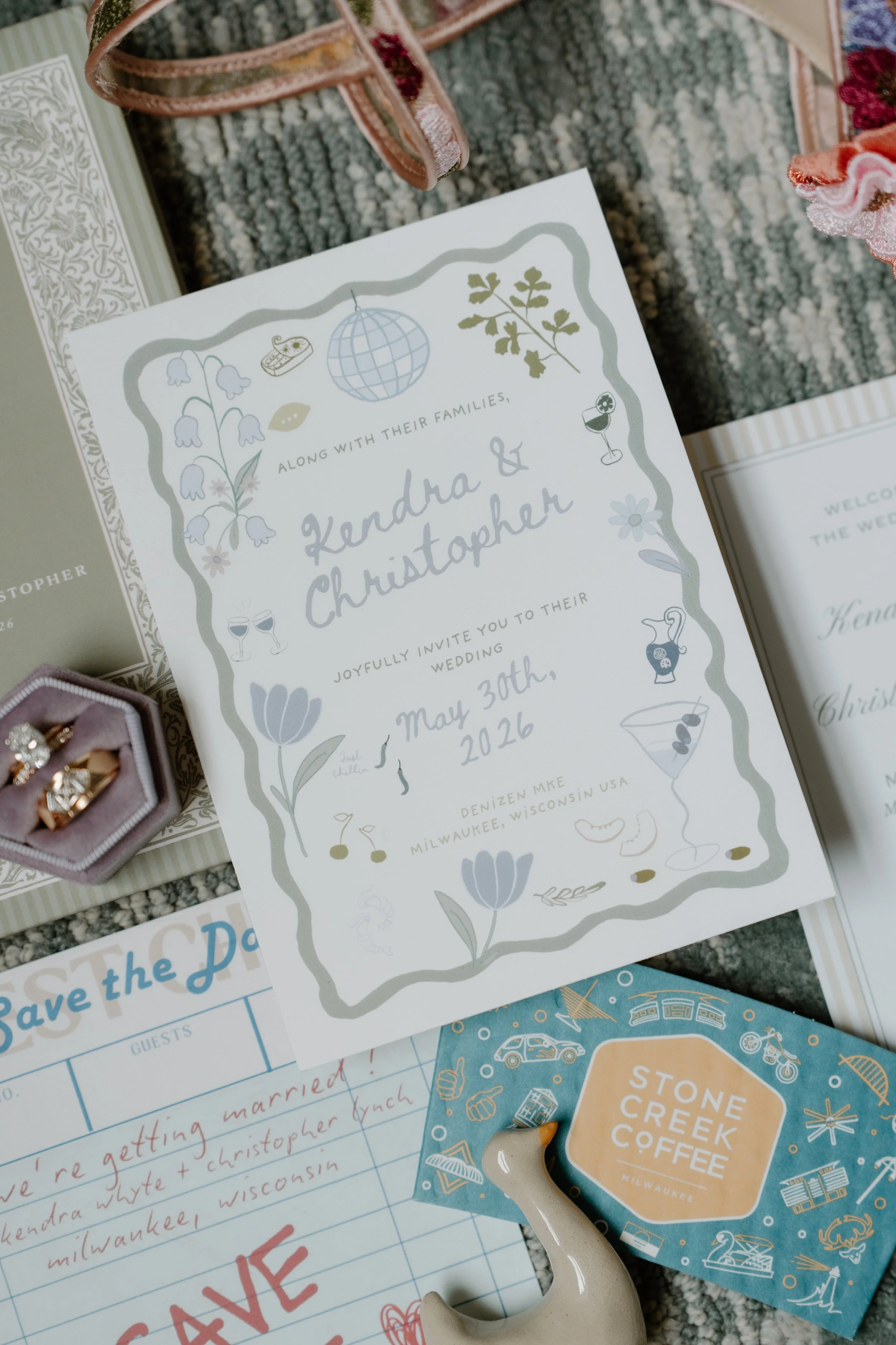 Wedding invitation for Kendra and Christopher with floral and drink illustrations, dated May 30th, 2026, on a textured grey carpet.