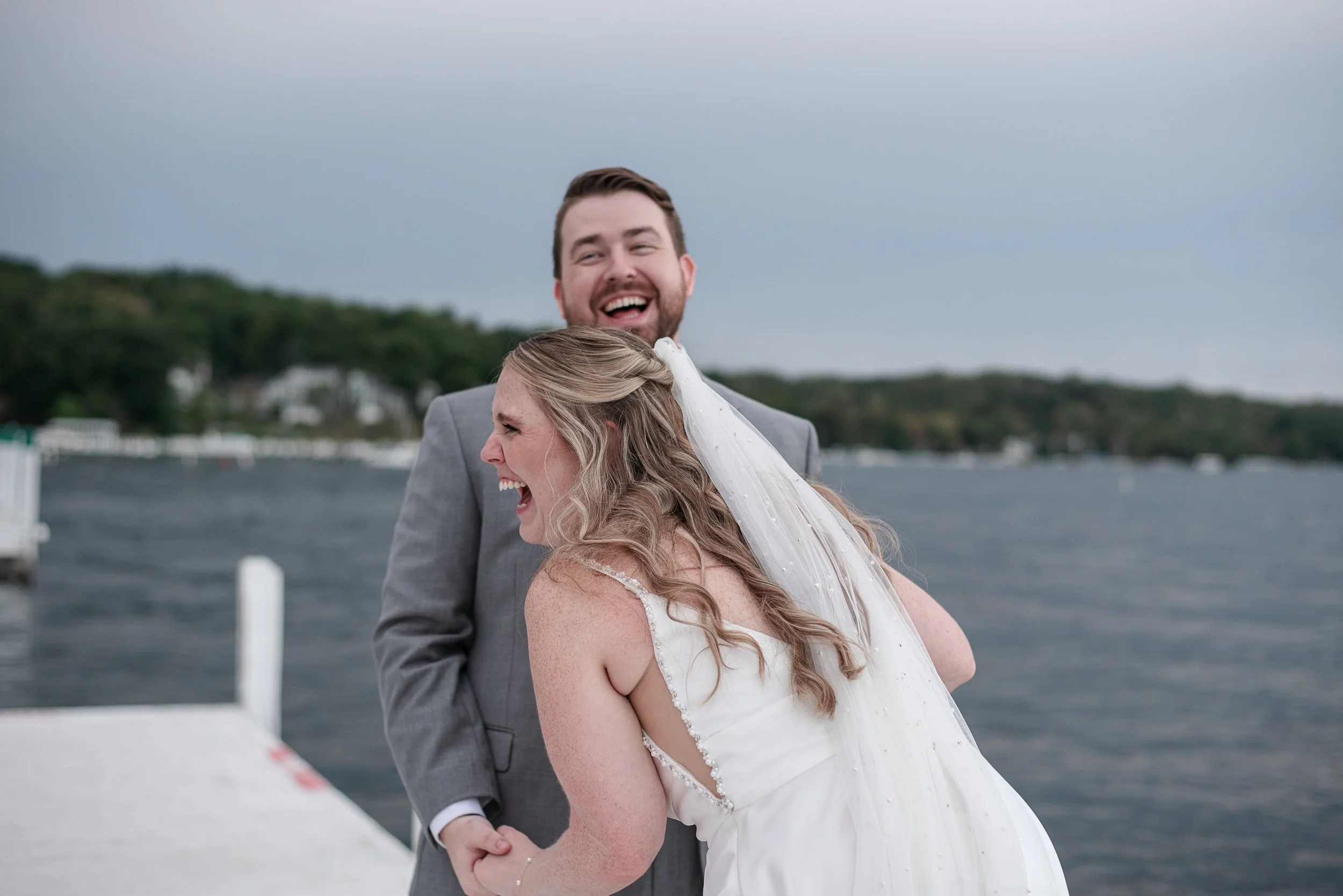 A happy bride and groom are laughing and smiling by the water, with a pier and some trees in the background.