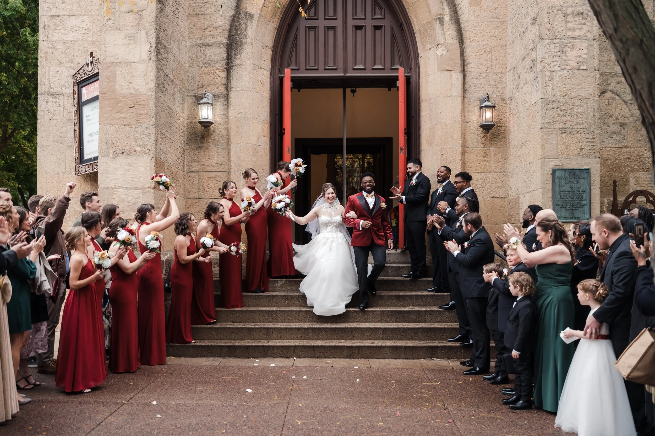 Bride and groom exiting a church surrounded by wedding guests cheering and taking photos, with bridesmaids and groomsmen on either side.