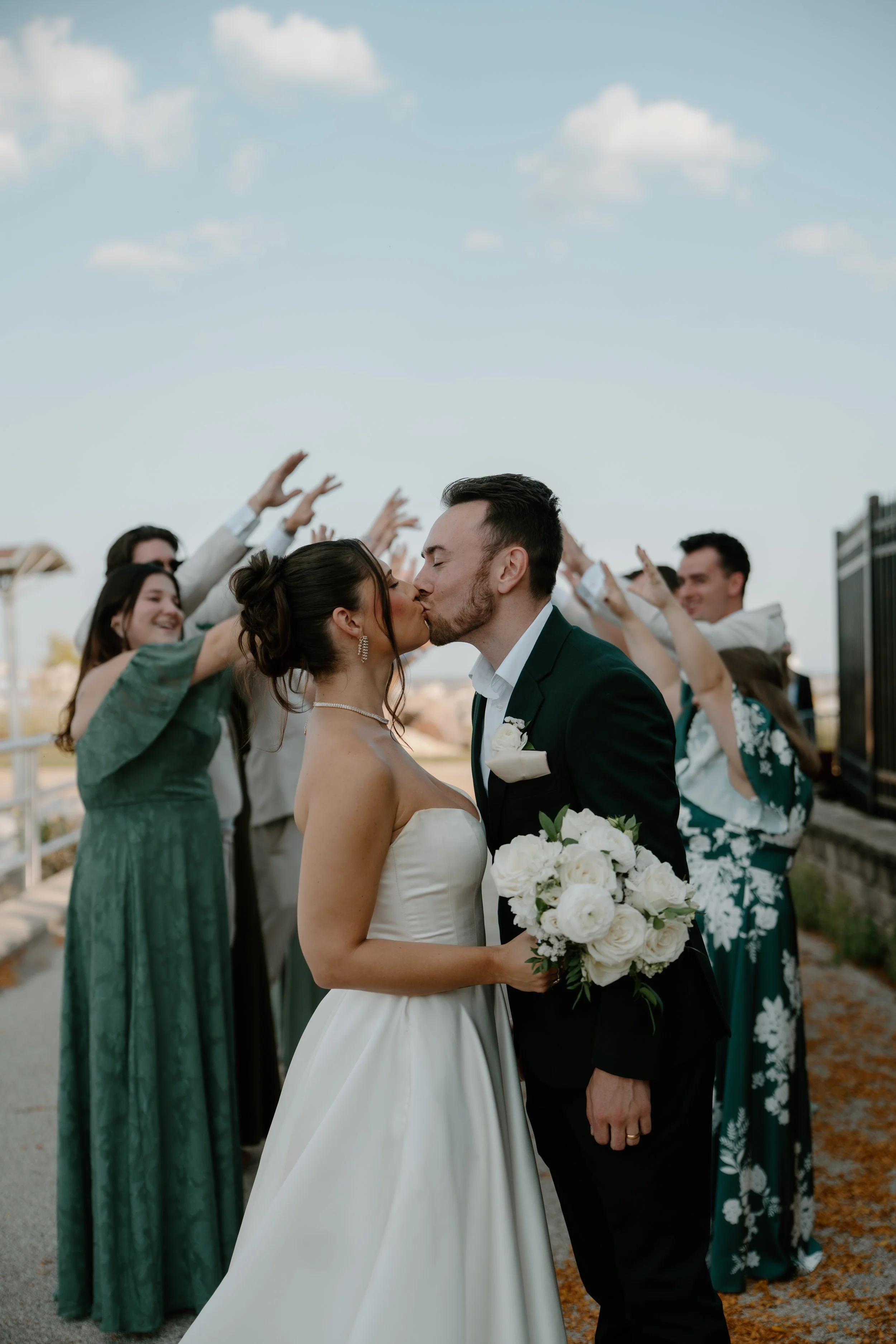 A bride and groom kissing at their wedding while friends cheer in the background, outdoors on a sunny day.