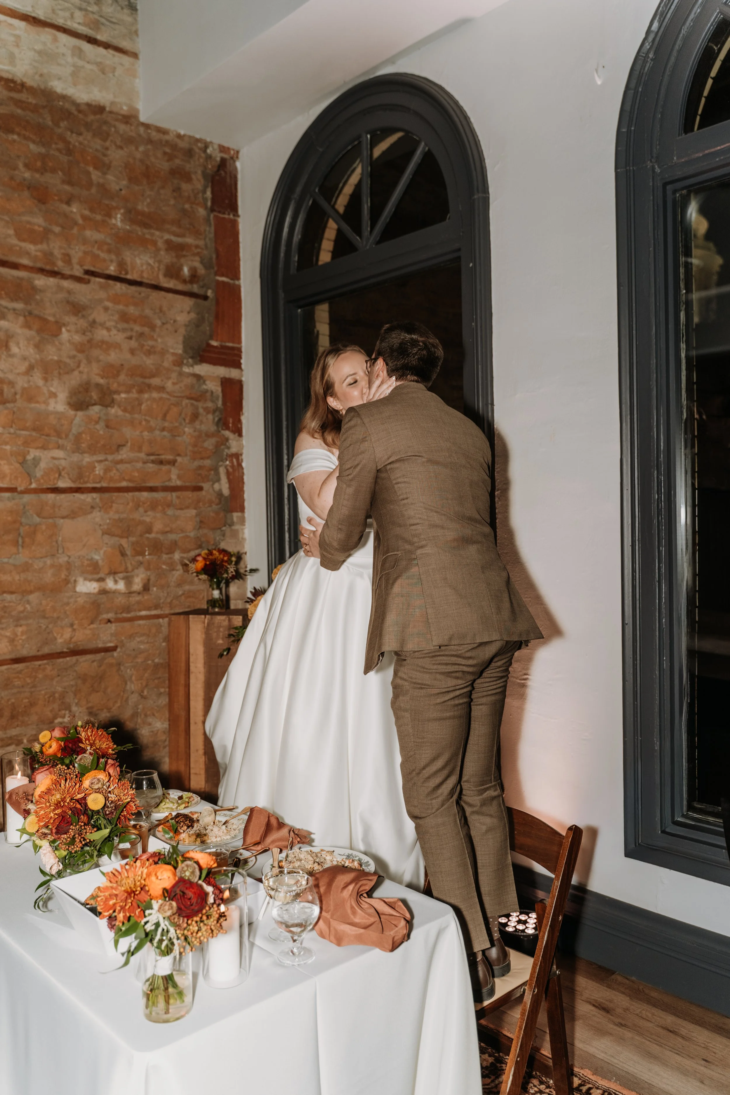 A couple sharing a kiss during a wedding reception, with a decorated table of flowers and food in the foreground, inside a rustic brick-walled venue.