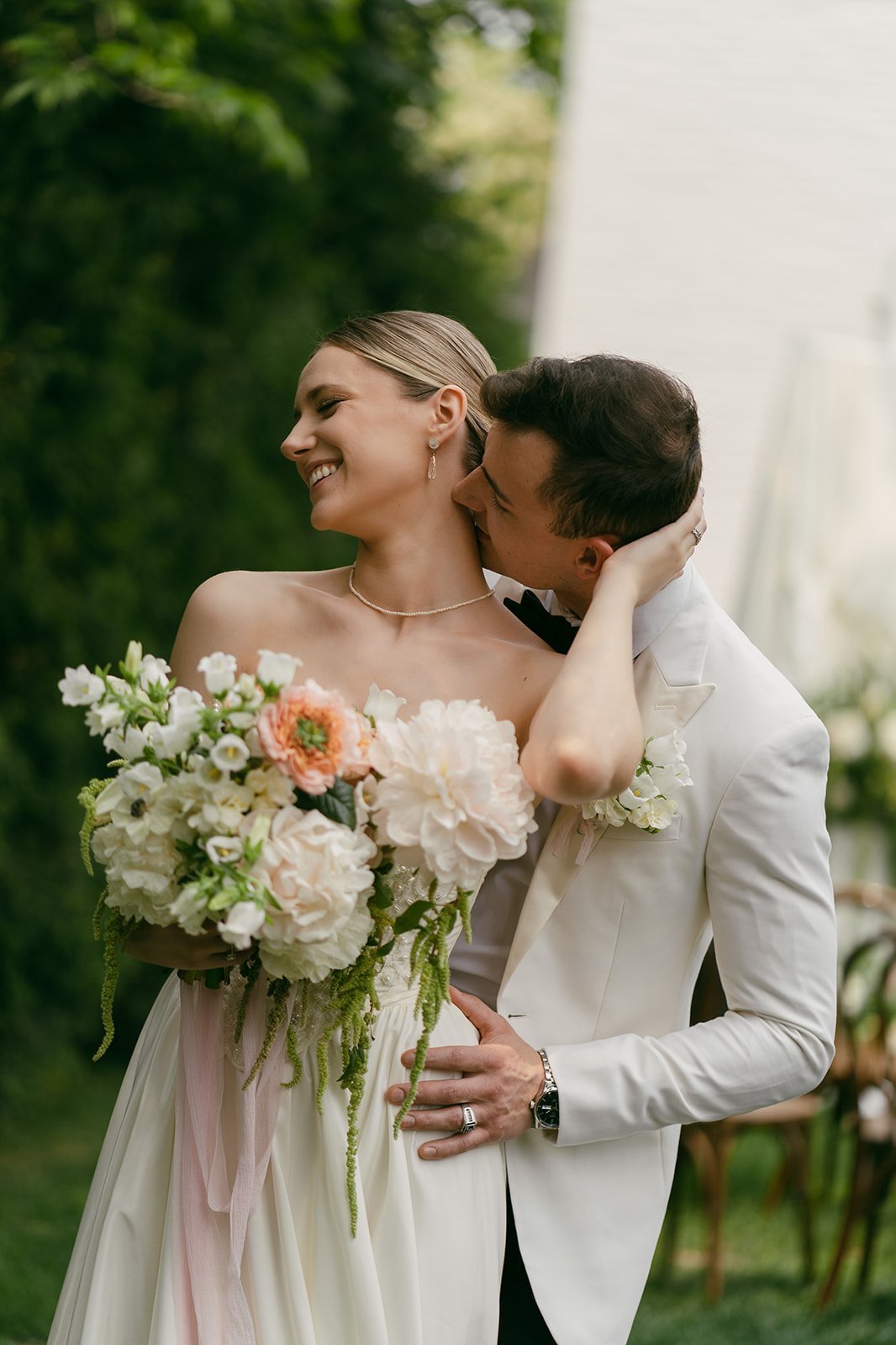 A newlywed couple sharing a kiss outdoors, with the groom in a white tuxedo and the bride holding a bouquet of white and pink flowers, surrounded by greenery.
