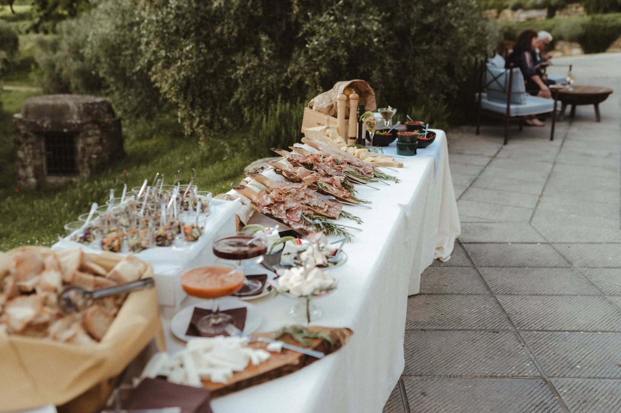 A long table outdoors with various foods, including cuts of meat, salads, bread, and drinks, set up for a gathering.
