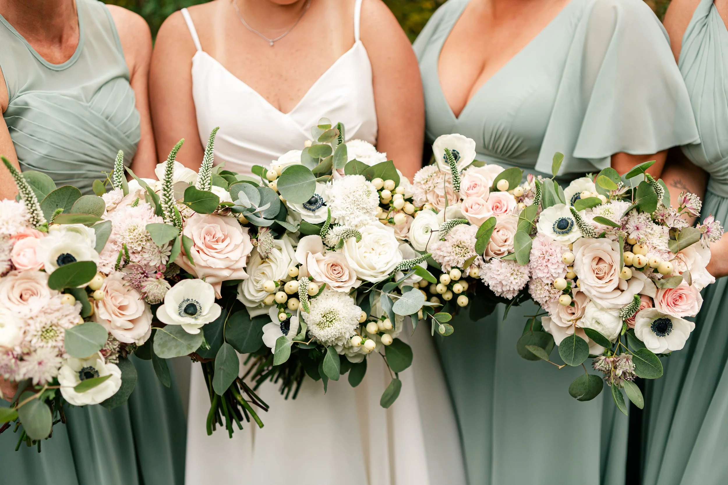A bride and bridesmaids holding a large bouquet of light pink and white roses, anemones, and greenery during a wedding ceremony.