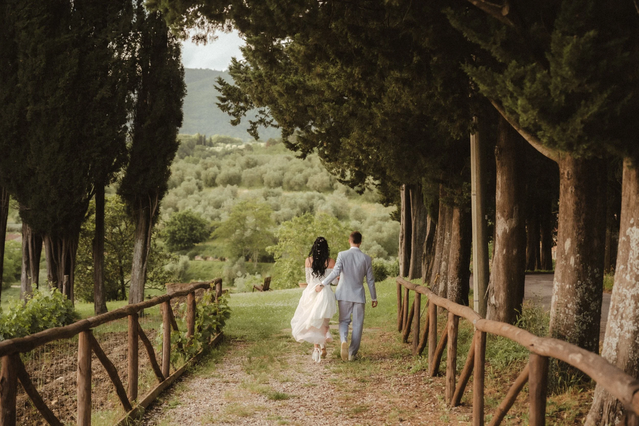 Couple dressed in wedding attire walking along a tree-lined path in a lush, green outdoor setting.