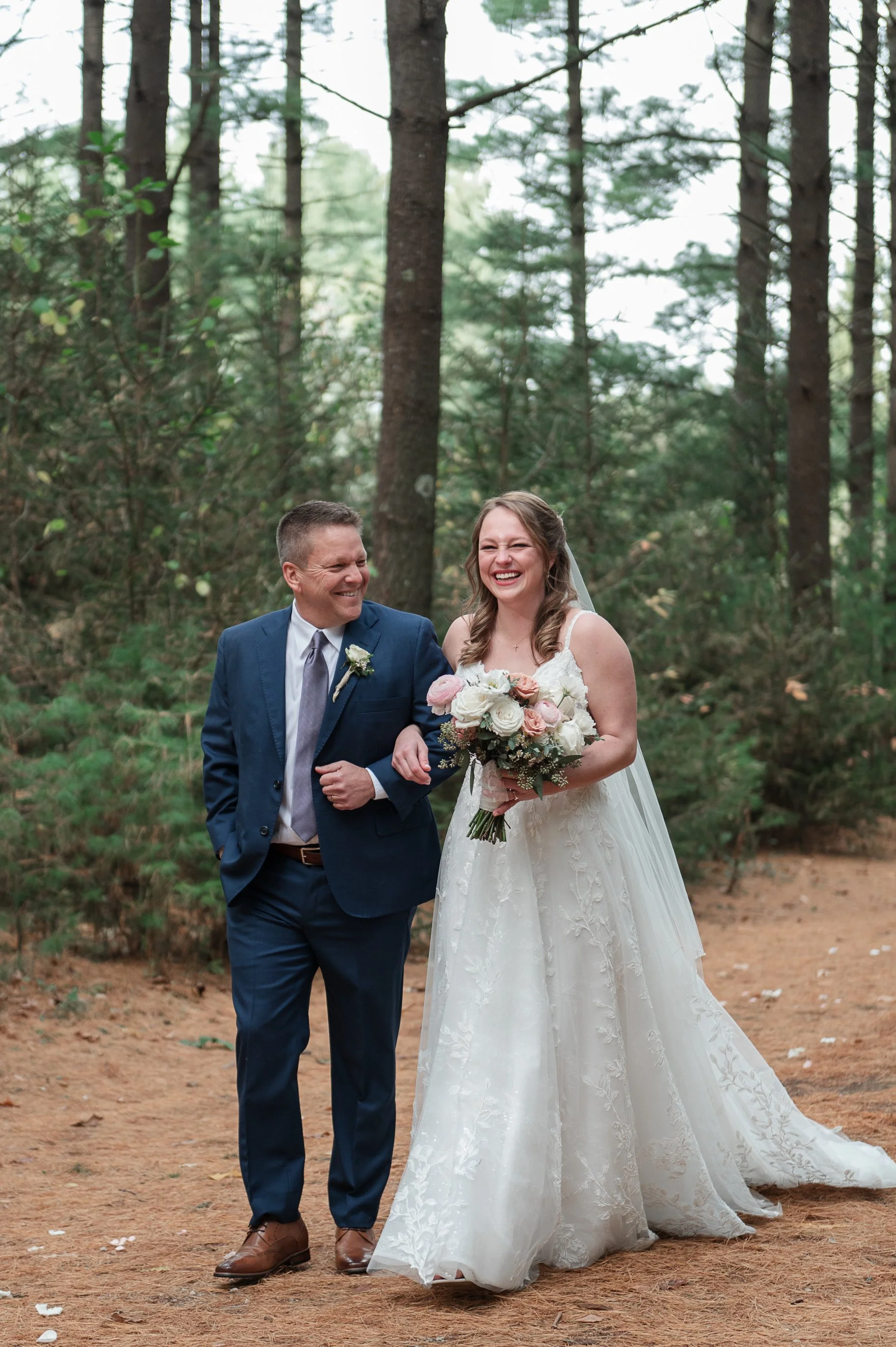 A bride walking down the woods with her father, both are smiling and the bride is holding a bouquet of pink and white flowers.