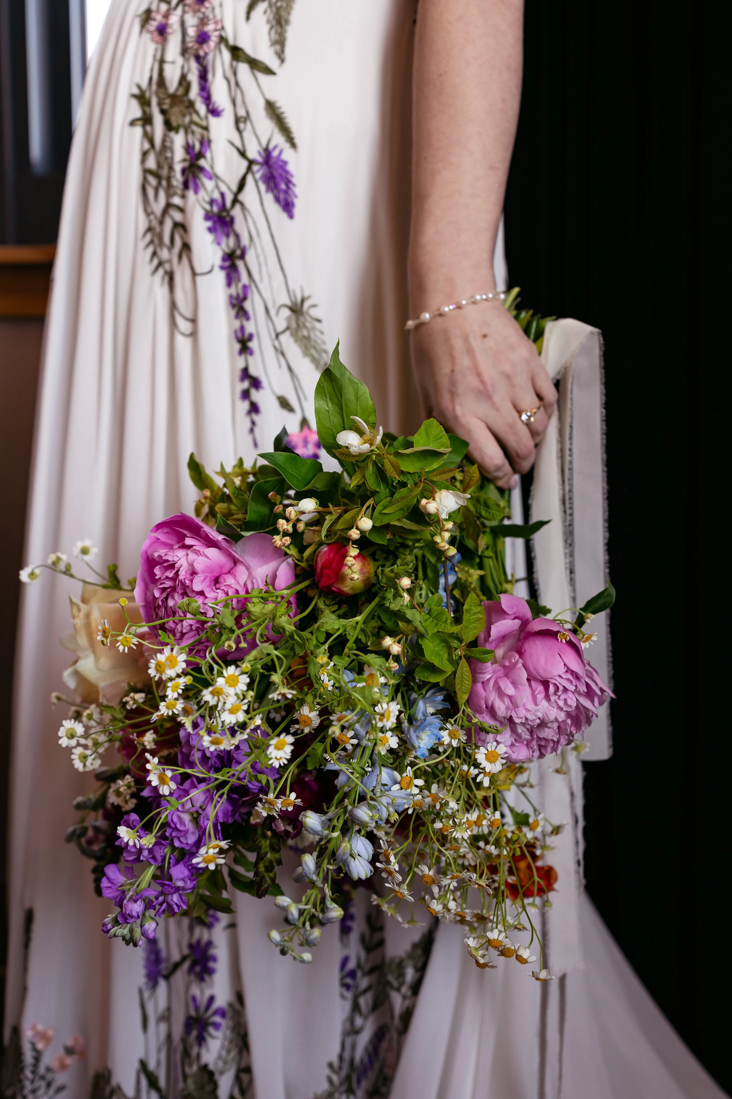 Close-up of a person wearing a white dress with purple floral embroidery, holding a bouquet of mixed flowers including pink peonies, white daisies, and purple and blue flowers, with a black background.